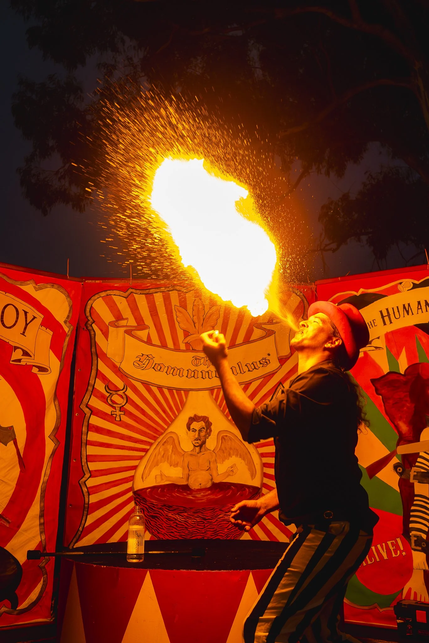 A circus performer dressed in striped pants and a red hat performs a fire-breathing act, creating a large burst of flames against a decorated carnival background.