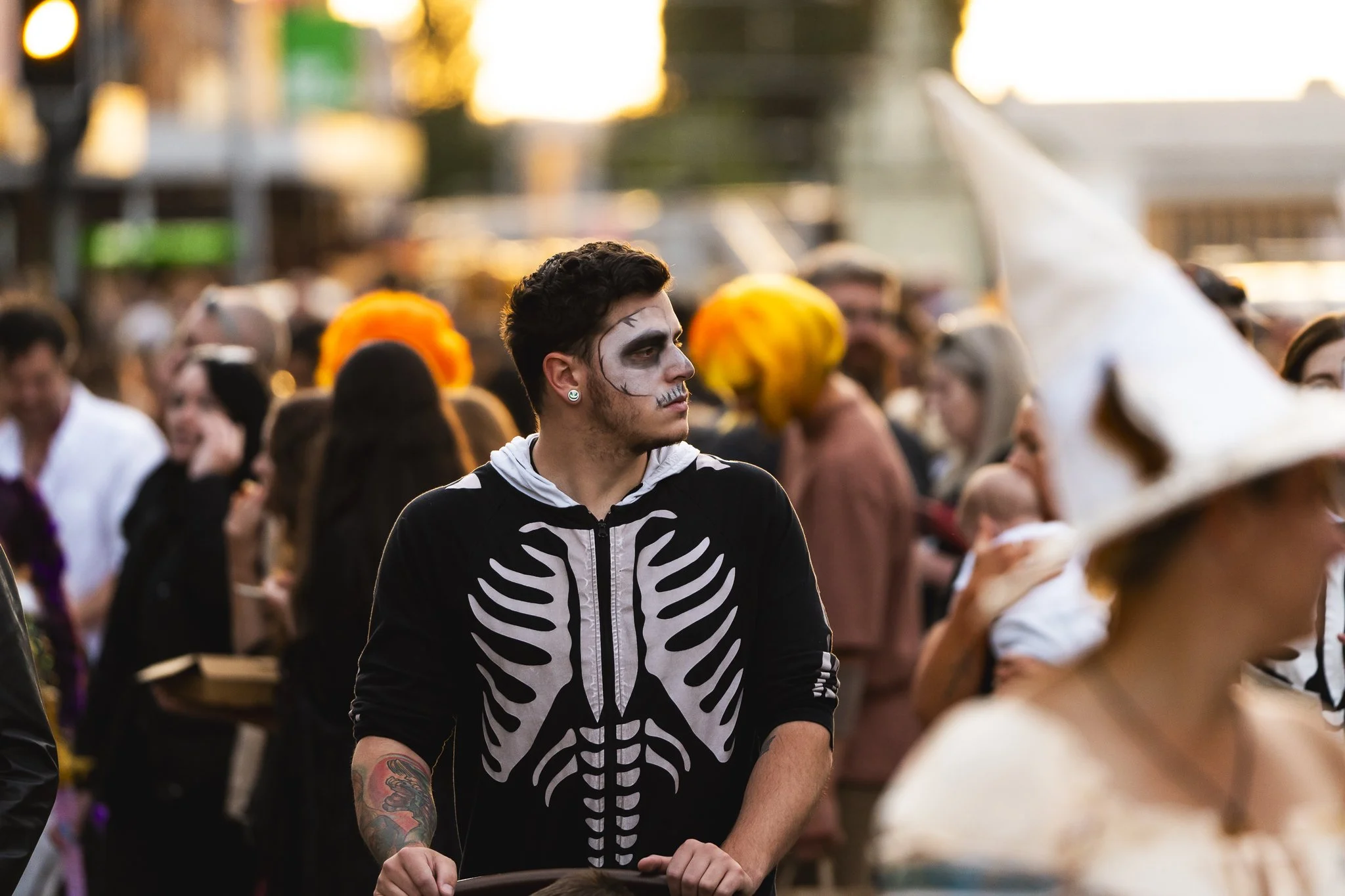 A young man dressed in skeleton-themed costume with face makeup and tattoos on his arm, standing in a crowd during a festive outdoor event, possibly Halloween or a costume parade, at sunset.