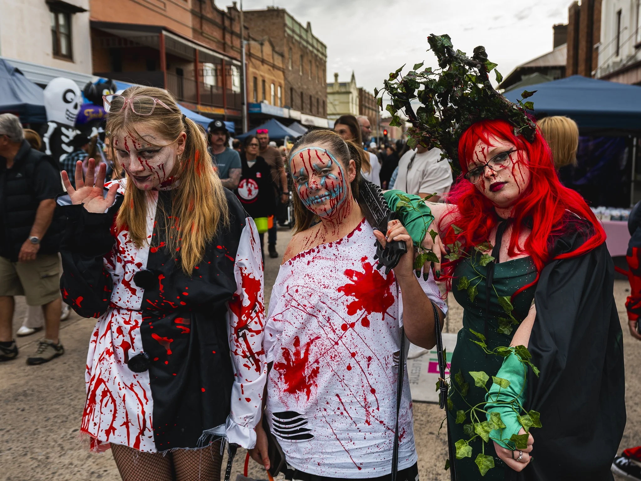 Three women dressed in horror-themed costumes at a crowded outdoor event. The woman on the left has long blonde hair, face makeup resembling a skull, and is wearing a blood-spattered black and white coat. The woman in the middle has face makeup with 