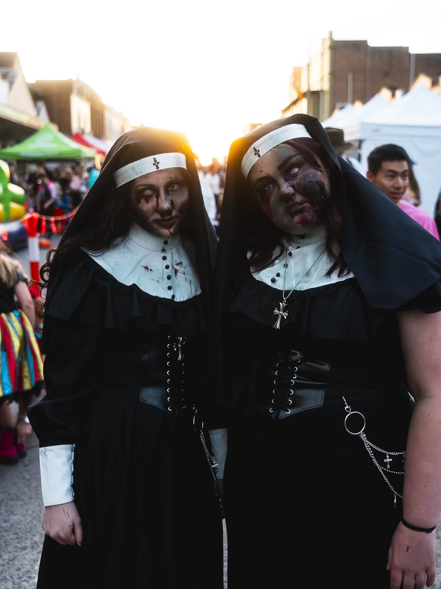 Two people dressed as sinister nuns with makeup resembling zombies, standing together at an outdoor festival or fair during sunset, with tents and other festival-goers in the background.