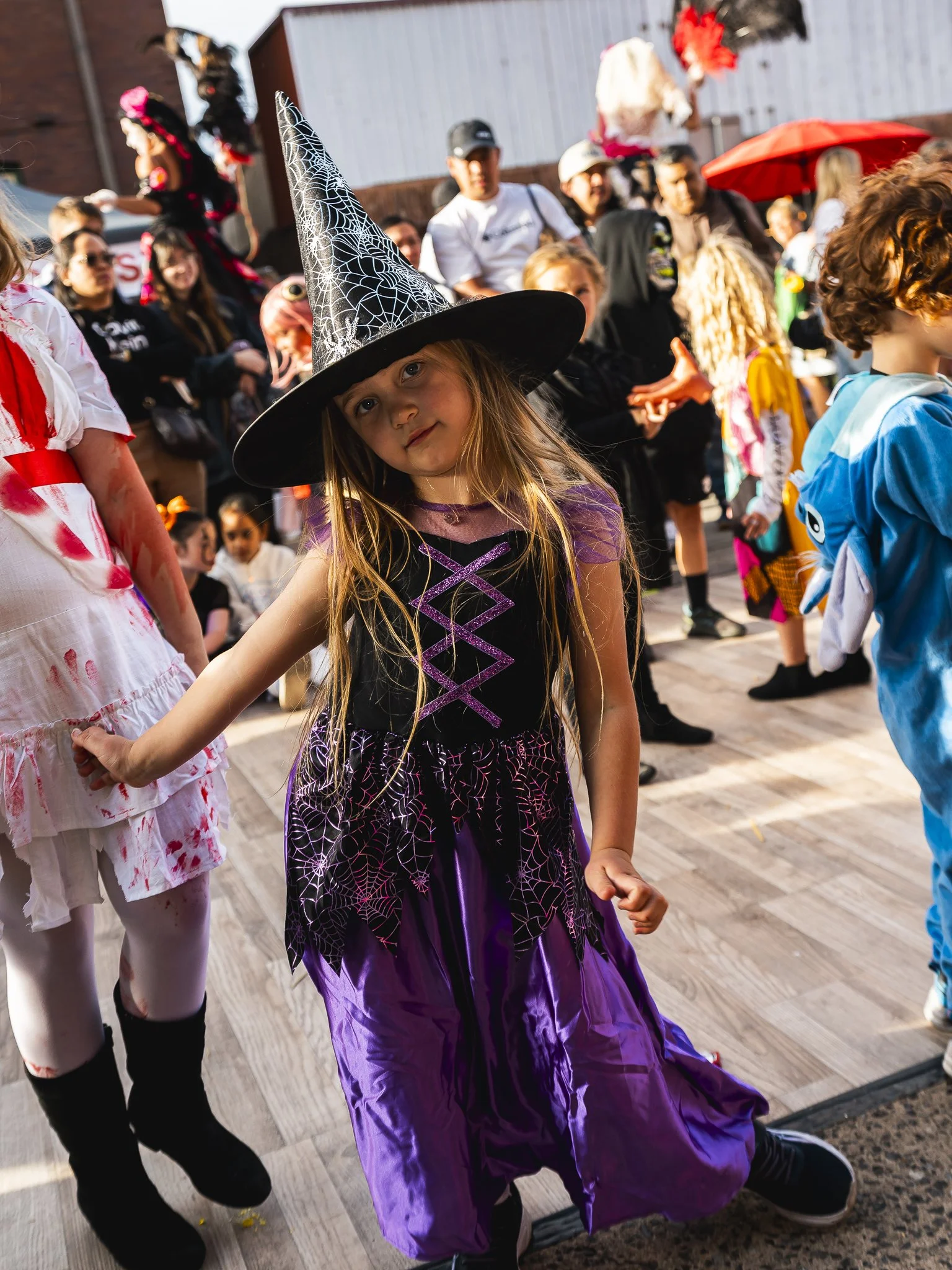 A young girl dressed as a witch in a purple and black costume with spider web designs, wearing a pointed witch hat, participating in a Halloween event with other children and adults dressed in costumes.