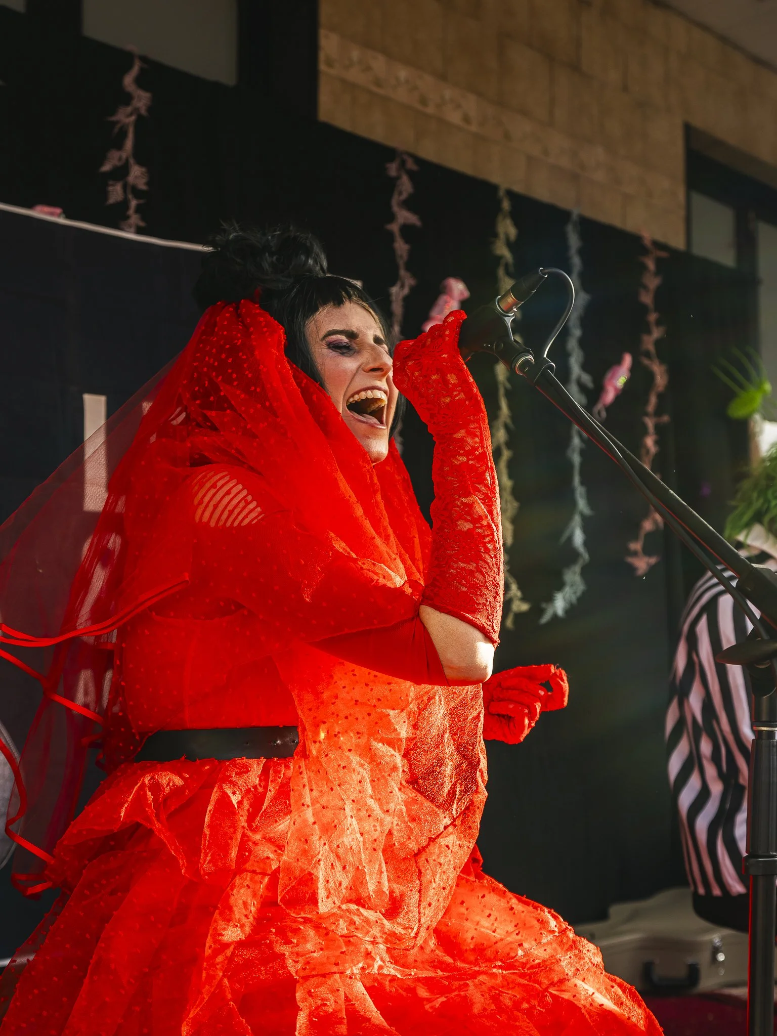 A woman in a vibrant red dress and lace gloves is singing passionately into a microphone during a performance.