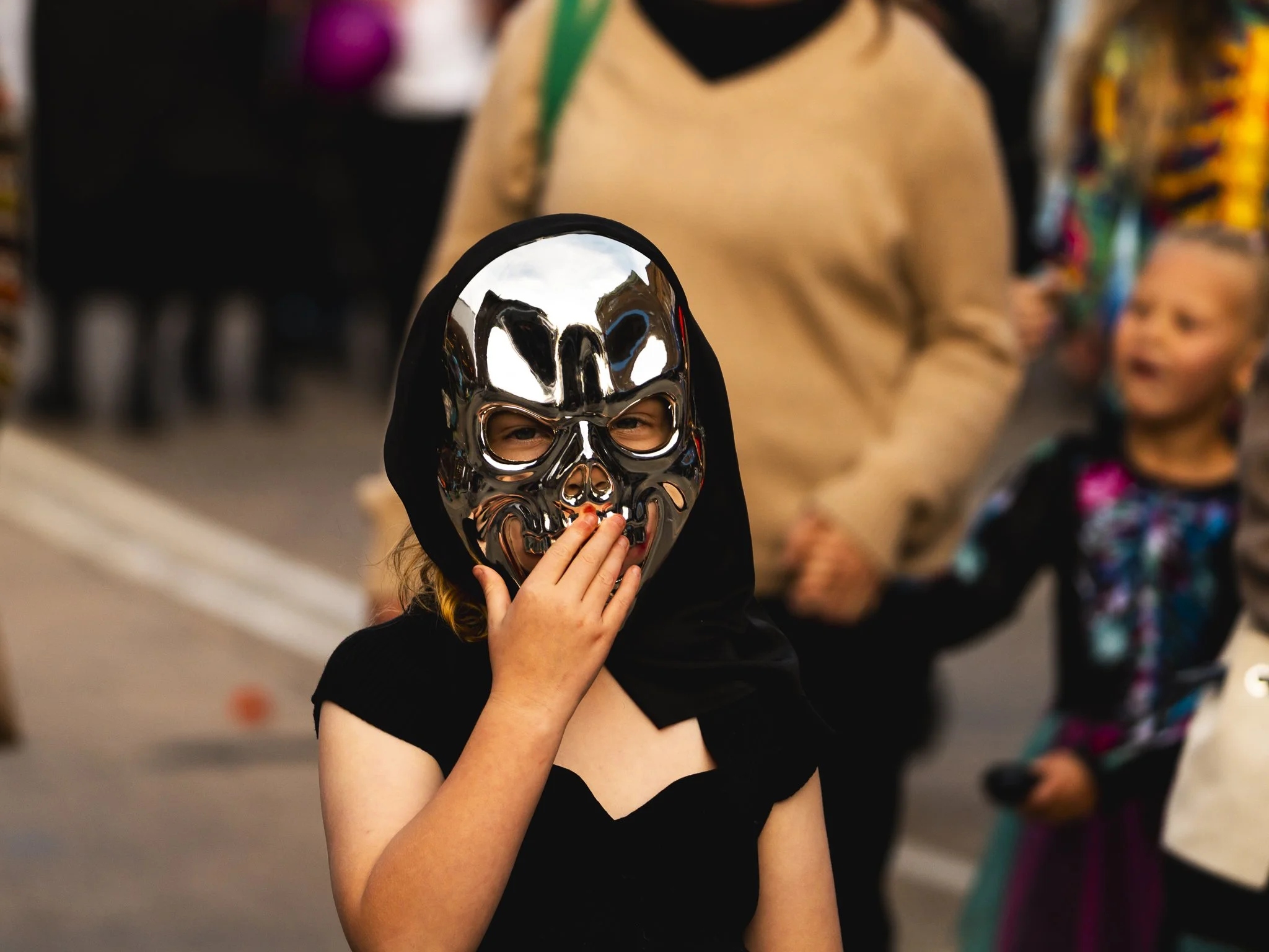 Child wearing a shiny metallic skull mask with black hood, holding hand to mouth, at a festive event with other children and adults in the background.