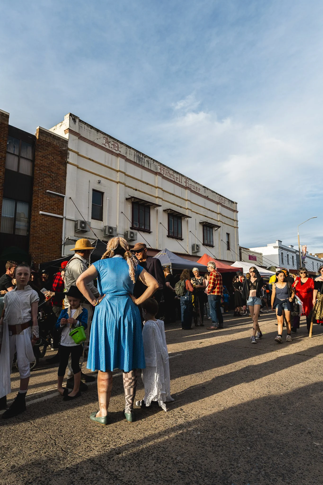 People walking and socializing at an outdoor marketplace or fair on a sunny day, with tents and an old building in the background.