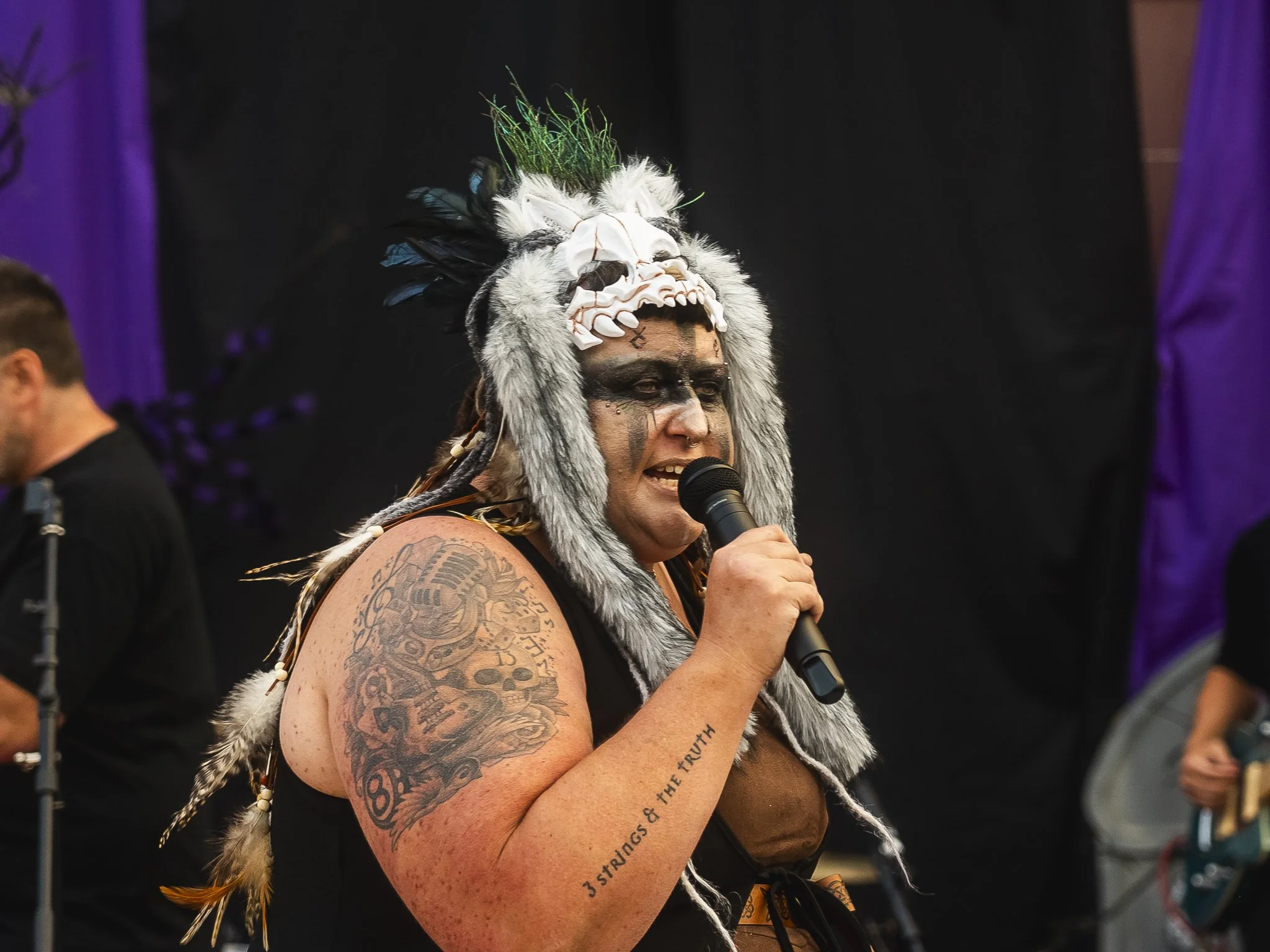 A woman wearing a tribal mask with animal features, feathers, and a headpiece, speaking into a microphone during a performance or event, with tattoos on her arm and dark makeup.