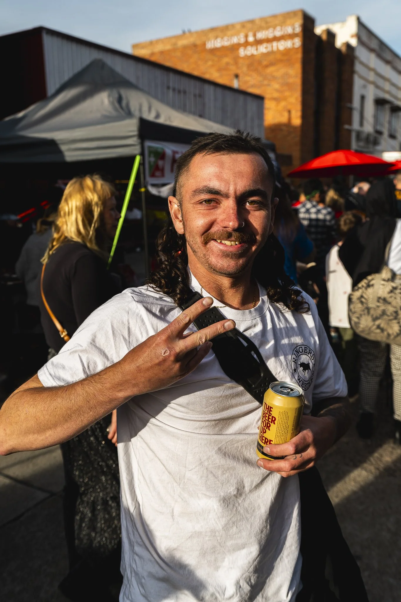 A man with dark hair and facial hair smiling at a crowded outdoor event, holding a can of beer and making a peace sign with his right hand, wearing a white t-shirt, with tents and people in the background.