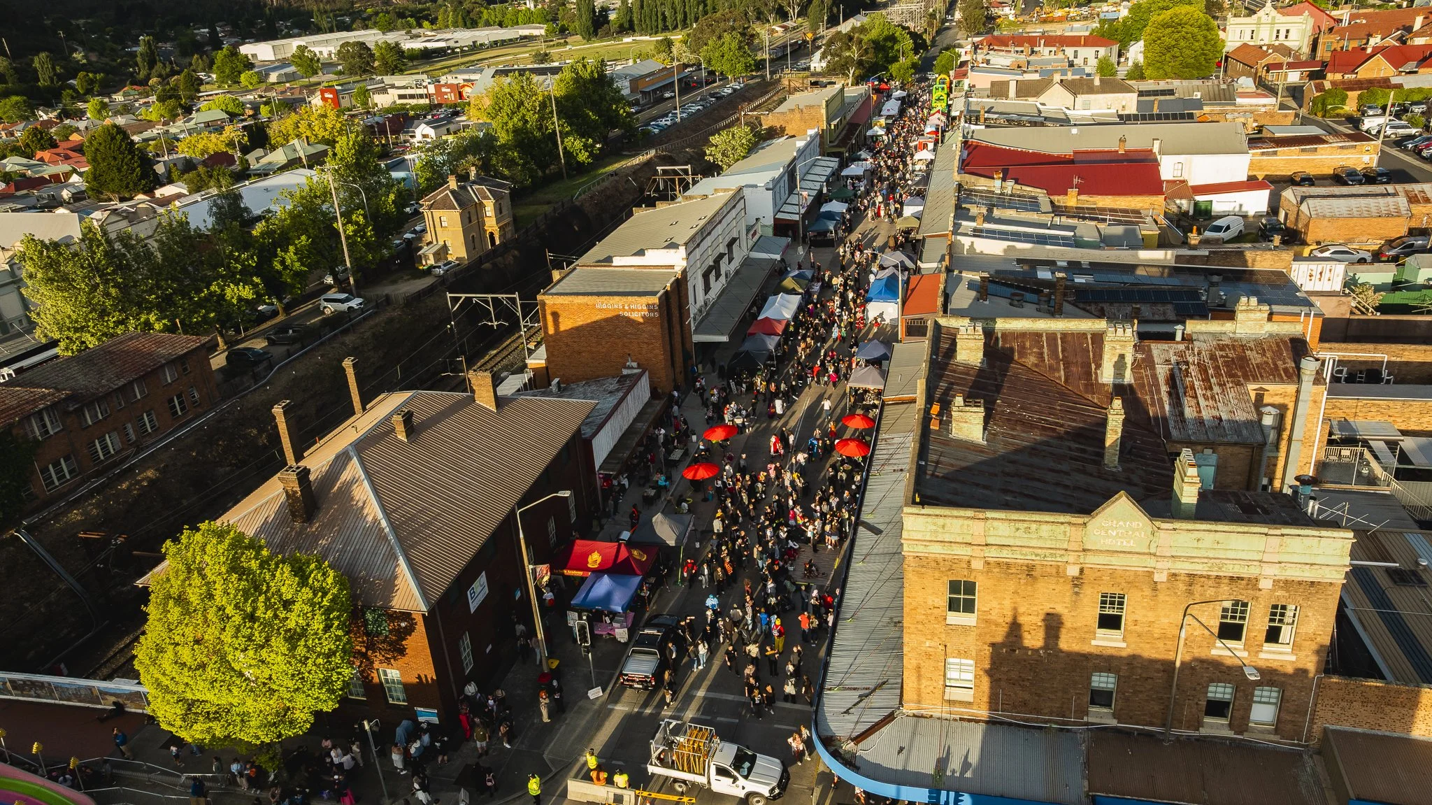 An aerial view of a busy outdoor street market with crowds of people, shops, and tents. Surrounding buildings include a hotel, age-old brick structures, and a green landscape with trees.