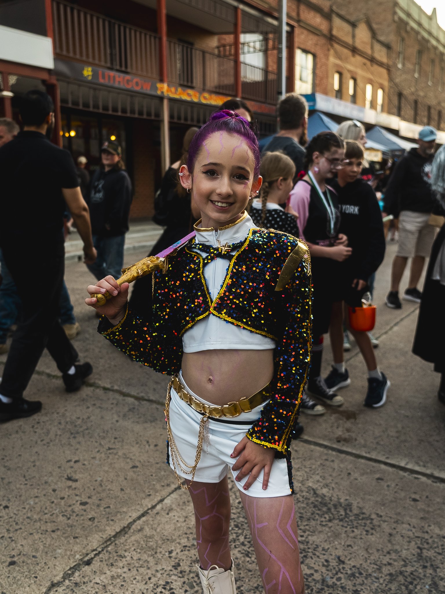 Young girl in a colorful, sequined costume with pink hair, standing on a sidewalk among a crowd at an outdoor event or festival, smiling and holding a prop sword.
