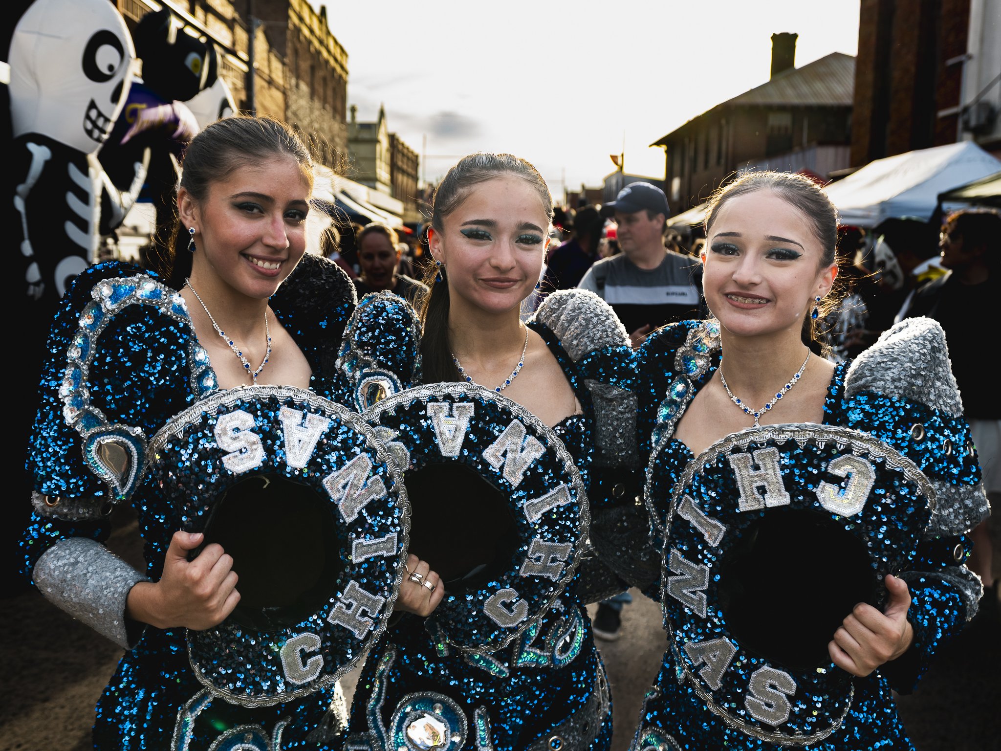 Three young women dressed in blue and silver sequined costumes with matching accessories, holding coiled sashes with their names, participating in a parade or festival, with spectators and street decorations in the background.