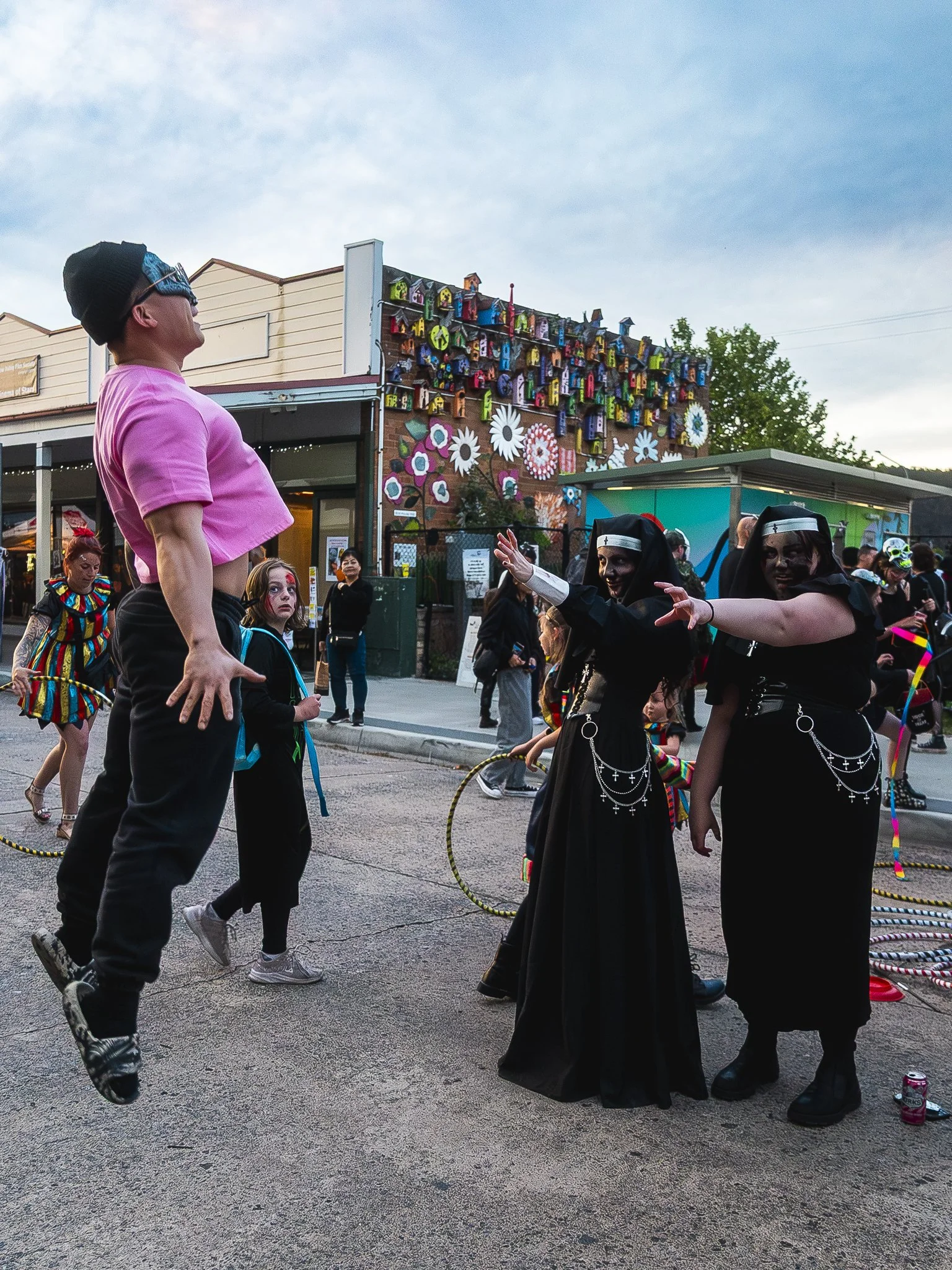 People dressed in costumes, including nuns and a person jumping, at a street event with colorful decorations and buildings in the background.