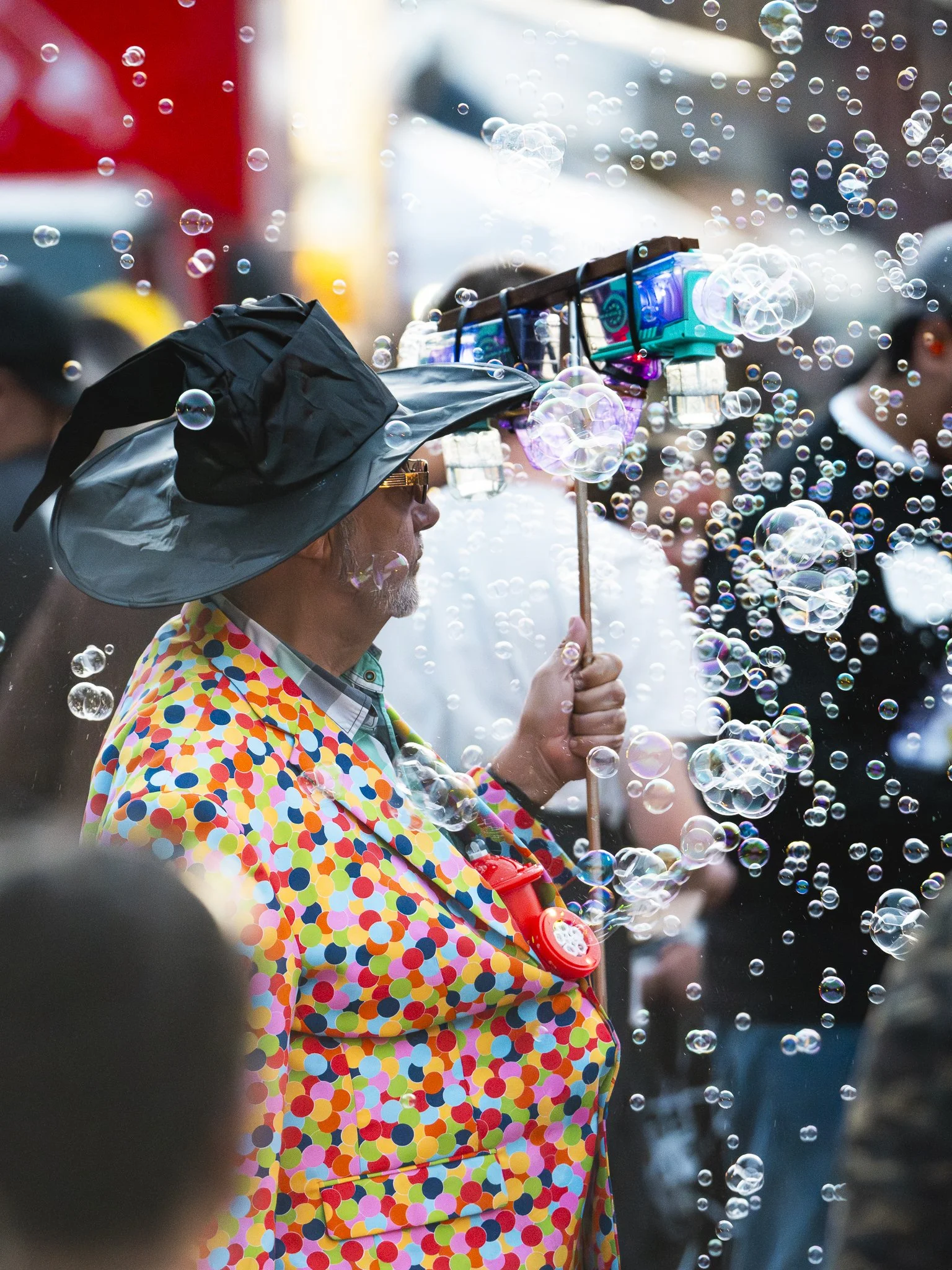 A man dressed in a polka dot colorful jacket and a large brimmed hat surrounded by bubbles at a public event.