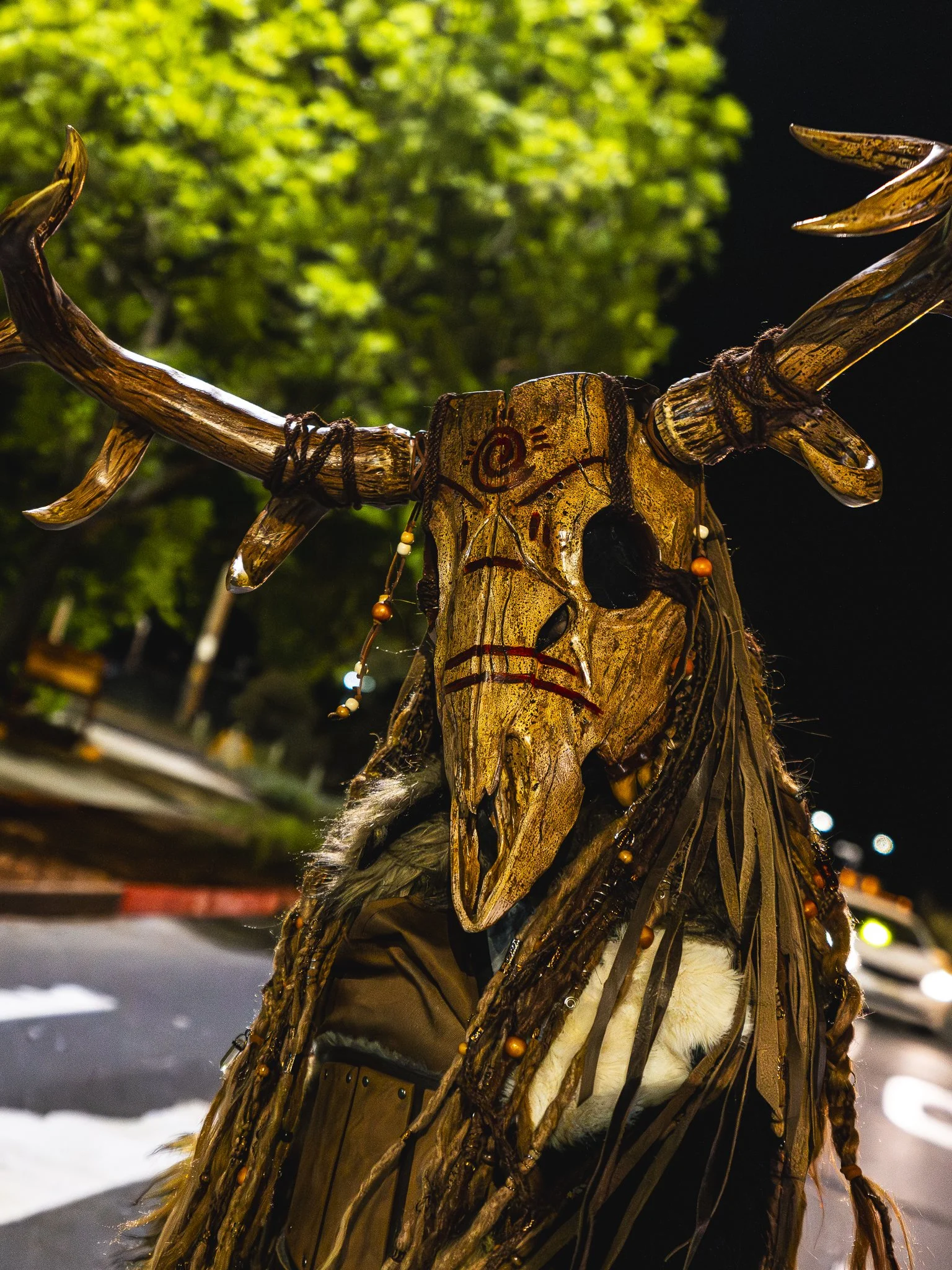 A person dressed in an elaborate costume resembling a tribal or fantasy character, with a large animal skull as a headdress, decorated with beads and feathers. The background shows trees and is lit at night.