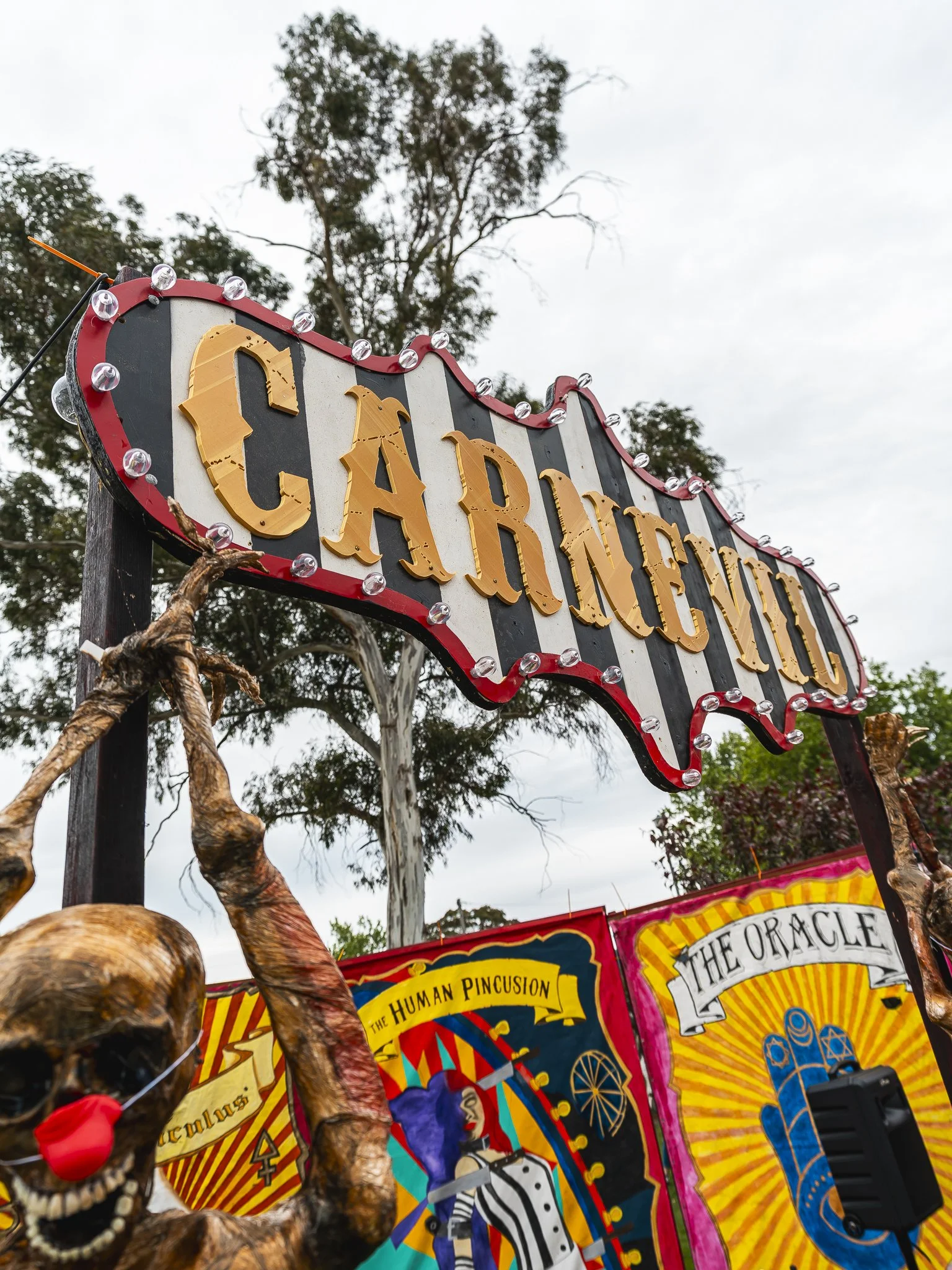 A carnival sign with black and white stripes, outlined in red, spelling out "CARNIVAL" in large yellow letters, decorated with small light bulbs. Below, there are colorful carnival-themed posters, including one titled "THE HUMAN PINCUSHION" with a cl