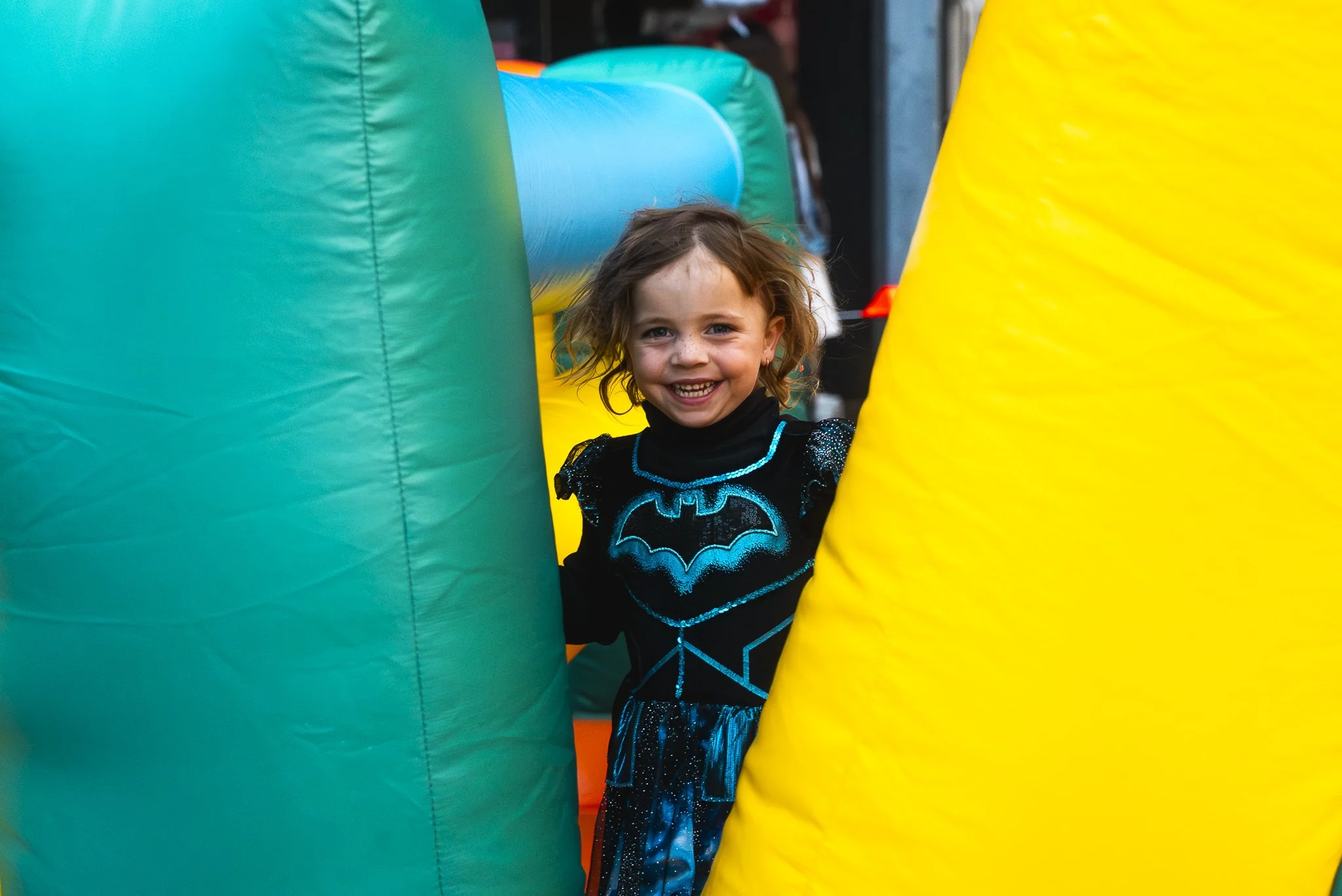 A young girl with curly hair and a big smile, wearing a black dress with a blue Batman logo, stands between large colorful inflatable structures in yellow, green, and blue.