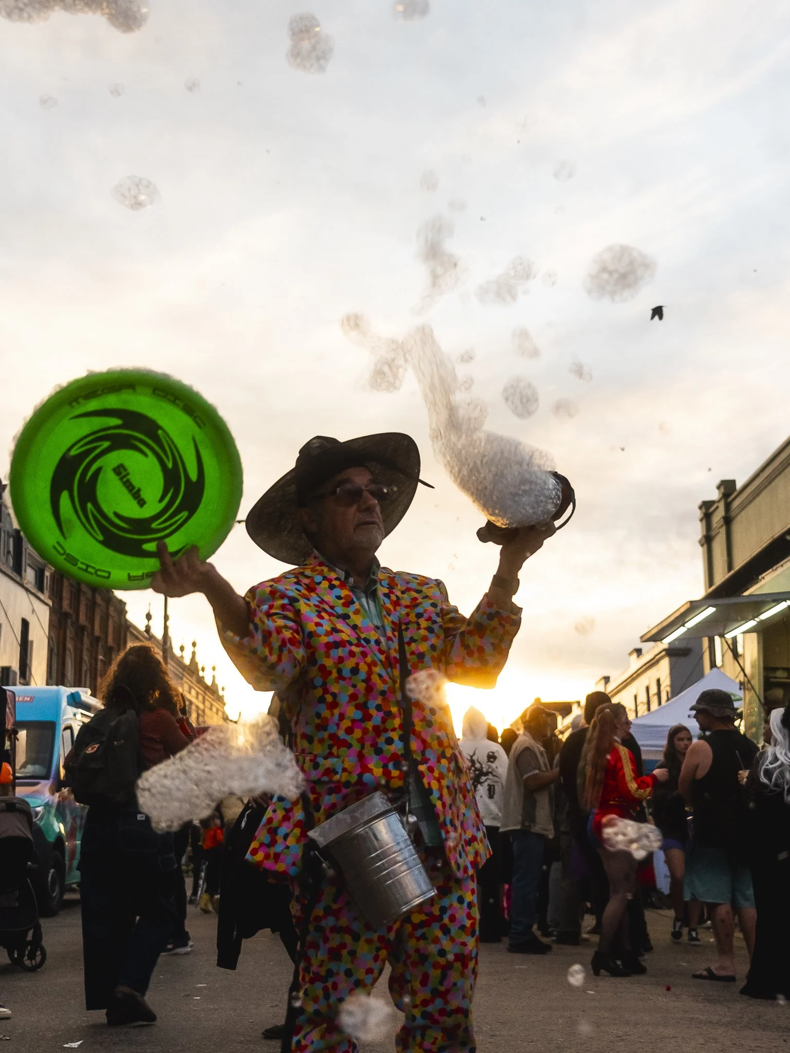 A street performer dressed in a colorful polka dot outfit, wearing a large hat, demonstrates a bubble-making act with a green bubble wand and a bubble machine in his hand, with a crowd and tents in the background during sunset.