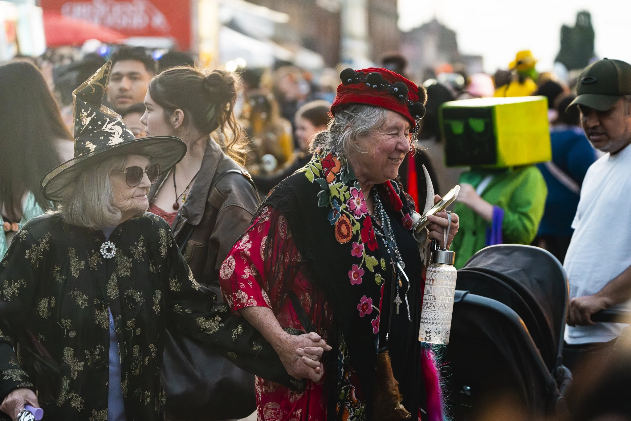 Two elderly women in colorful clothing and accessories hold hands amidst a crowd at an outdoor event, one with a witch hat and sunglasses, the other with a red headscarf and floral garments.