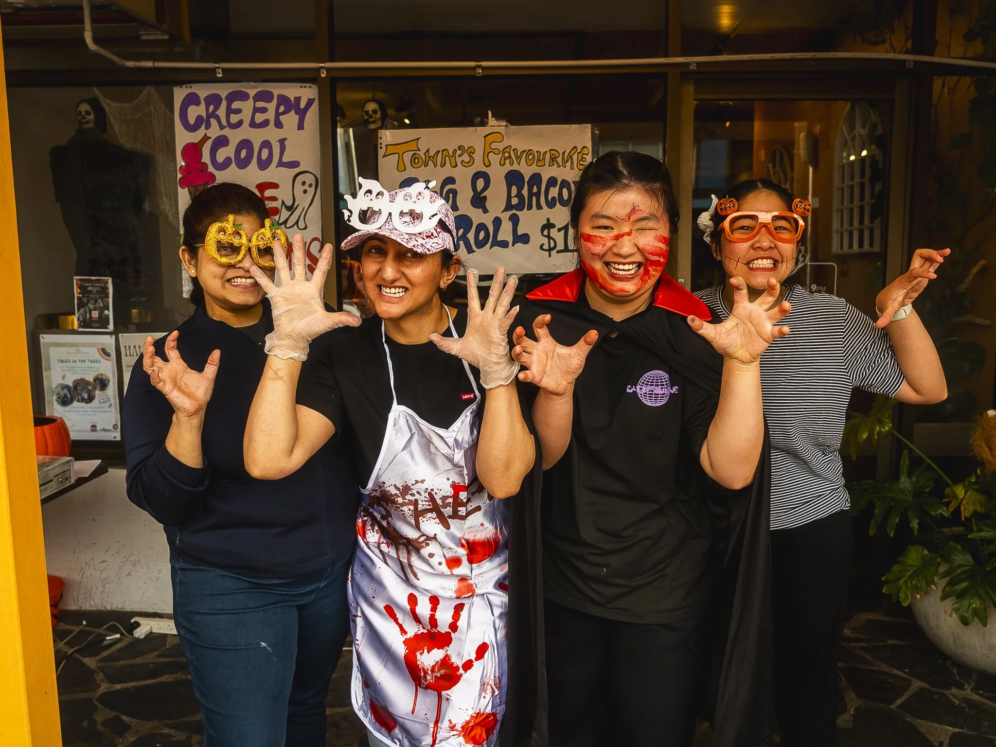Four women dressed in Halloween costumes, smiling and posing with their hands raised in front of a Halloween-themed stand with signs that read 'Creepy Cool' and 'John's Favourites Egg & Bacon Roll.' The women are wearing novelty glasses, a blood-spla