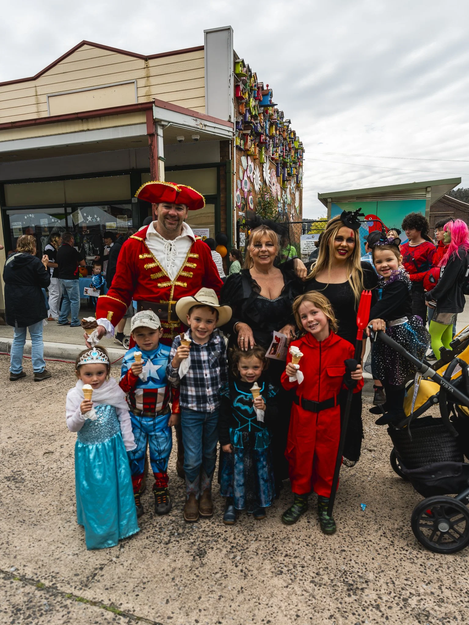 People dressed in costumes at a festival, including children in princess, superhero, and cowboy outfits, group photo with adults, some holding ice cream cones, outdoors in front of a building decorated with colorful flowers and sculptures.