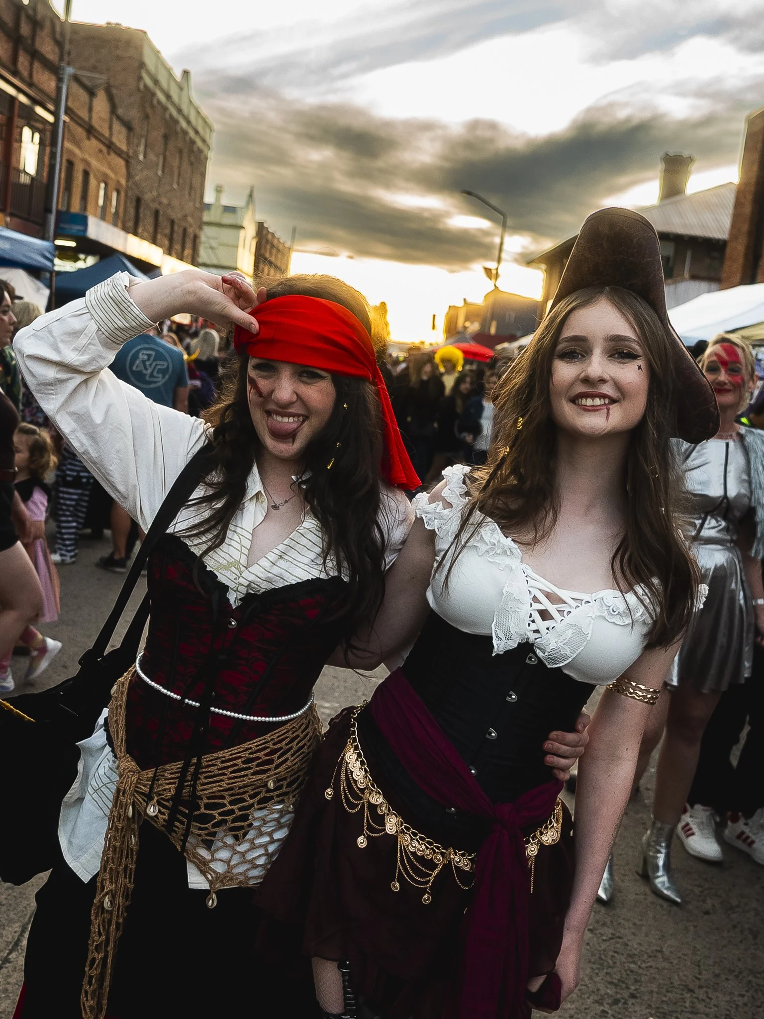Two women in pirate costumes at a festival during sunset, surrounded by people and market stalls.