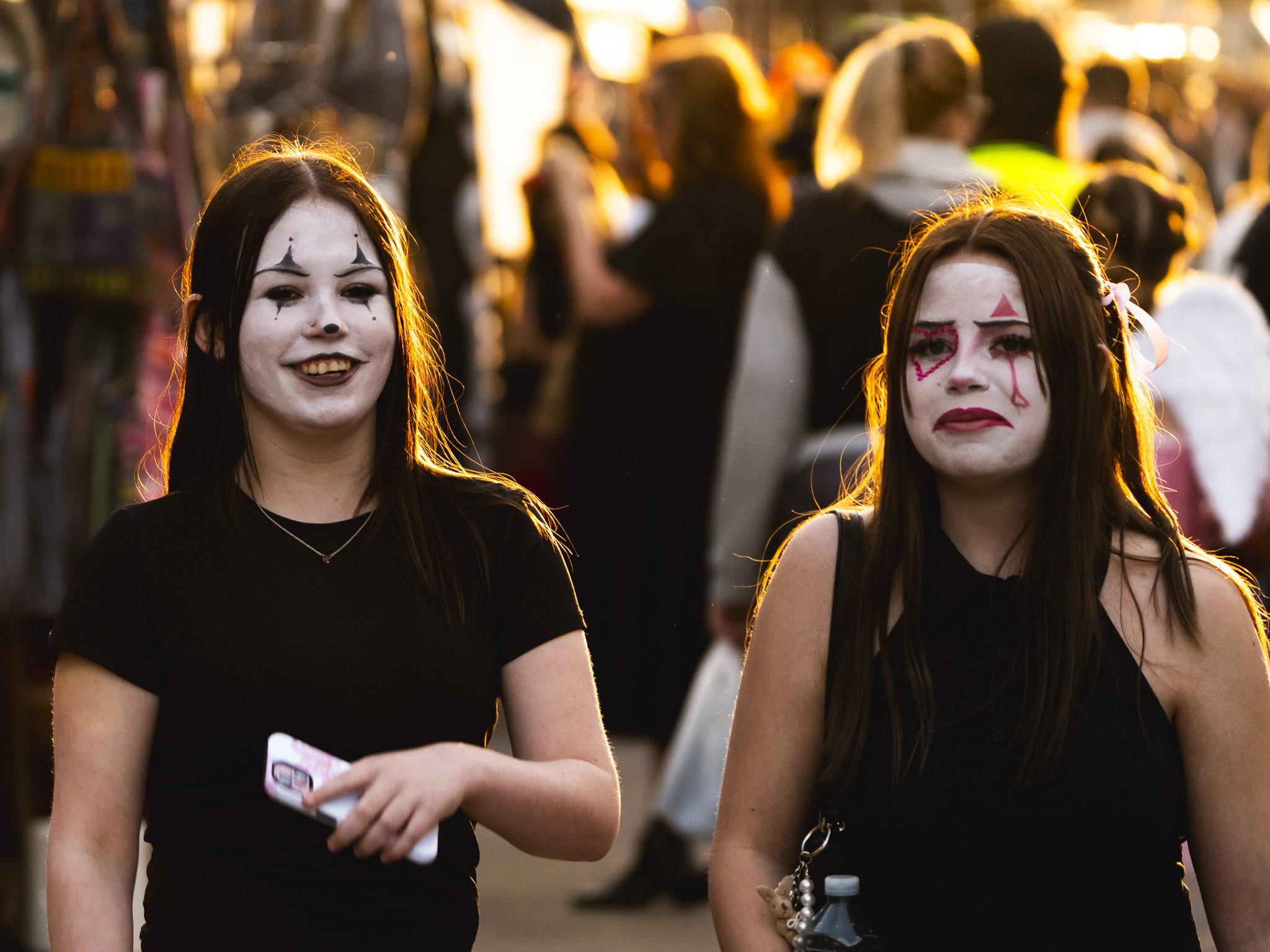 Two young women dressed as clowns with white face paint, dark hair, and dark clothing, standing in a crowded outdoor setting during sunset, with some people in the background.