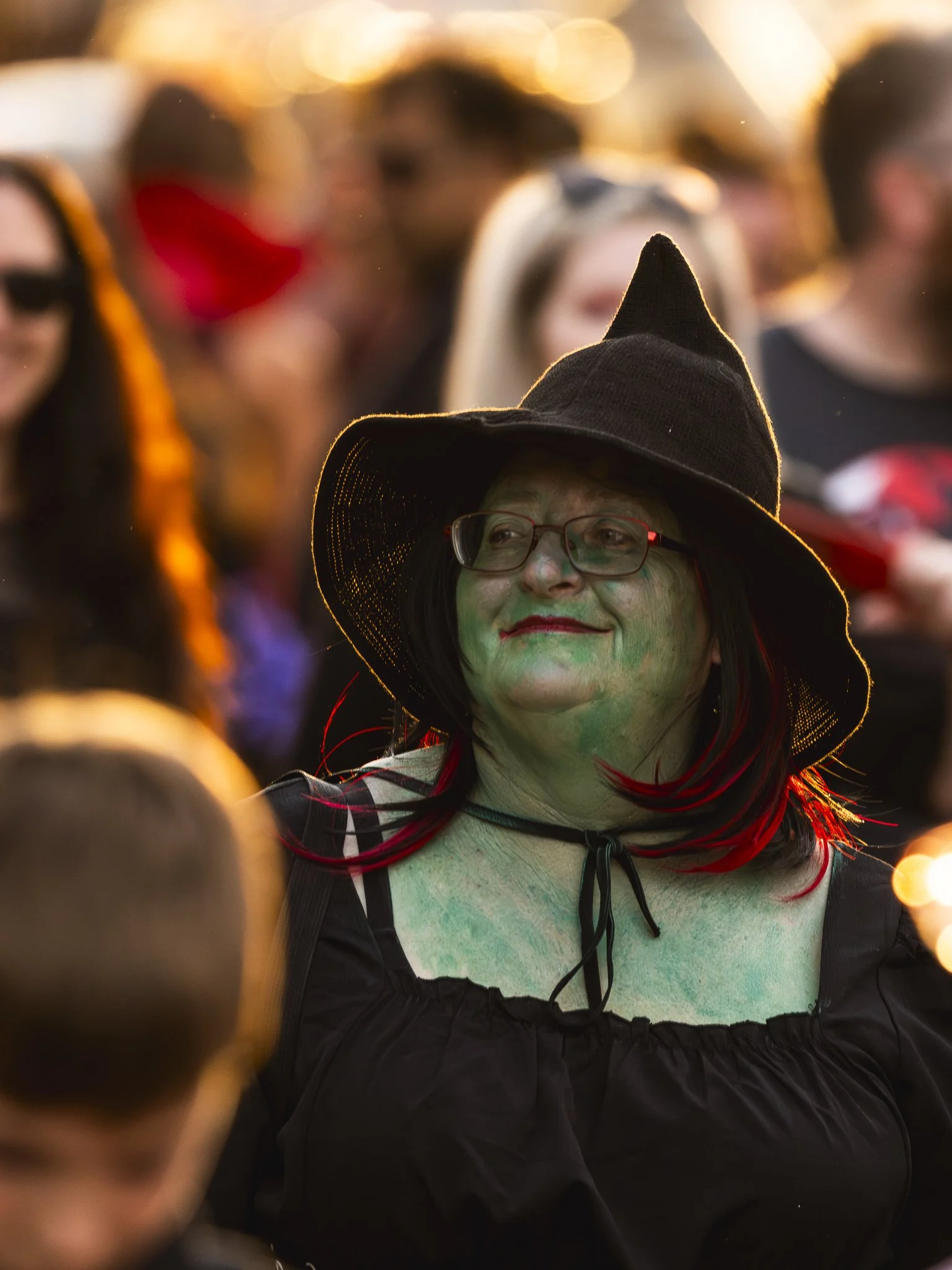 A person dressed as a witch with green face makeup, red hair, glasses, and a black hat, attending a costume event at sunset.