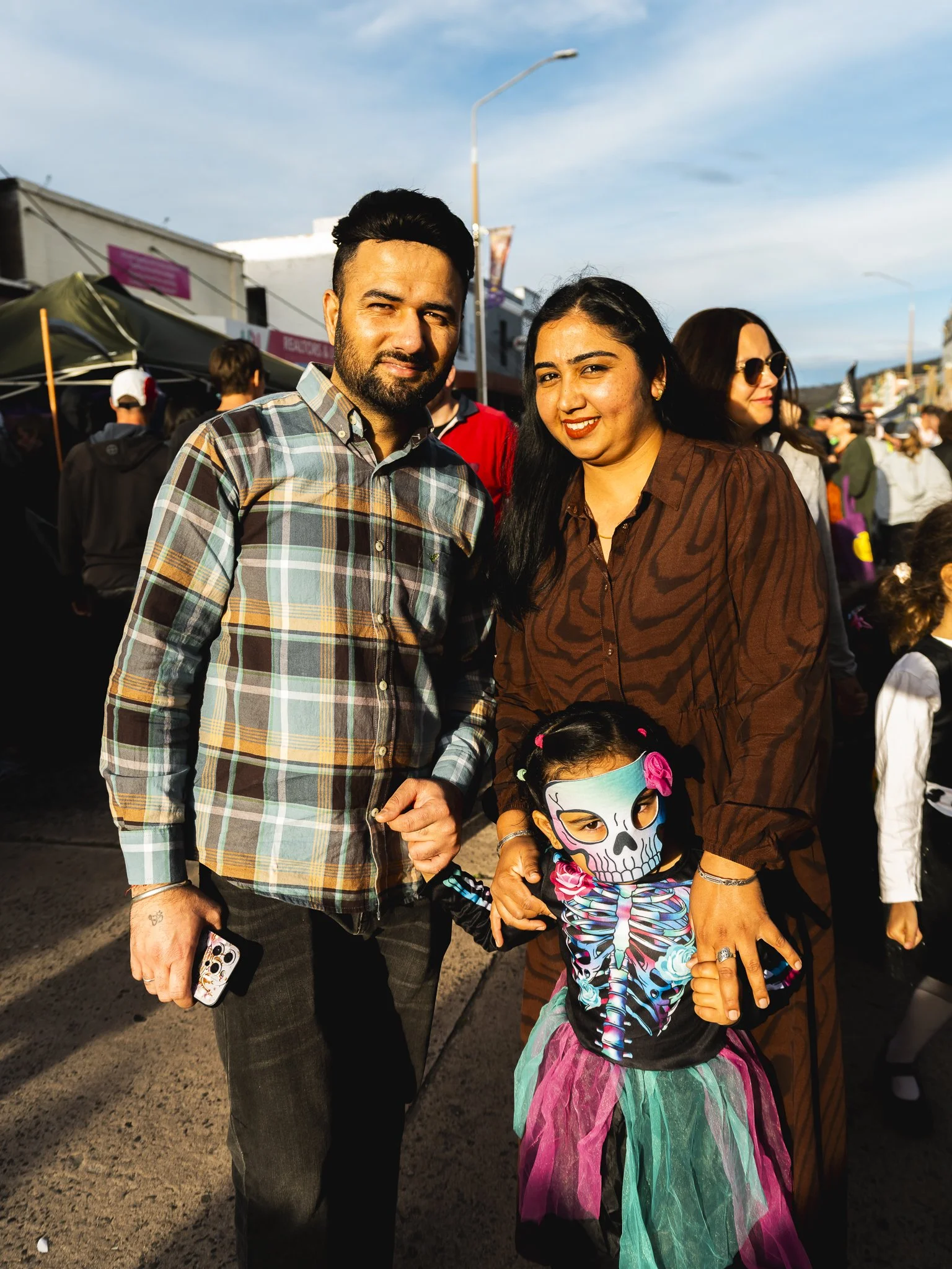 A family of three at an outdoor event during daytime, with a man, woman, and young girl dressed in colorful costumes, including a skeleton-themed dress for the girl with face paint, smiling and holding hands.