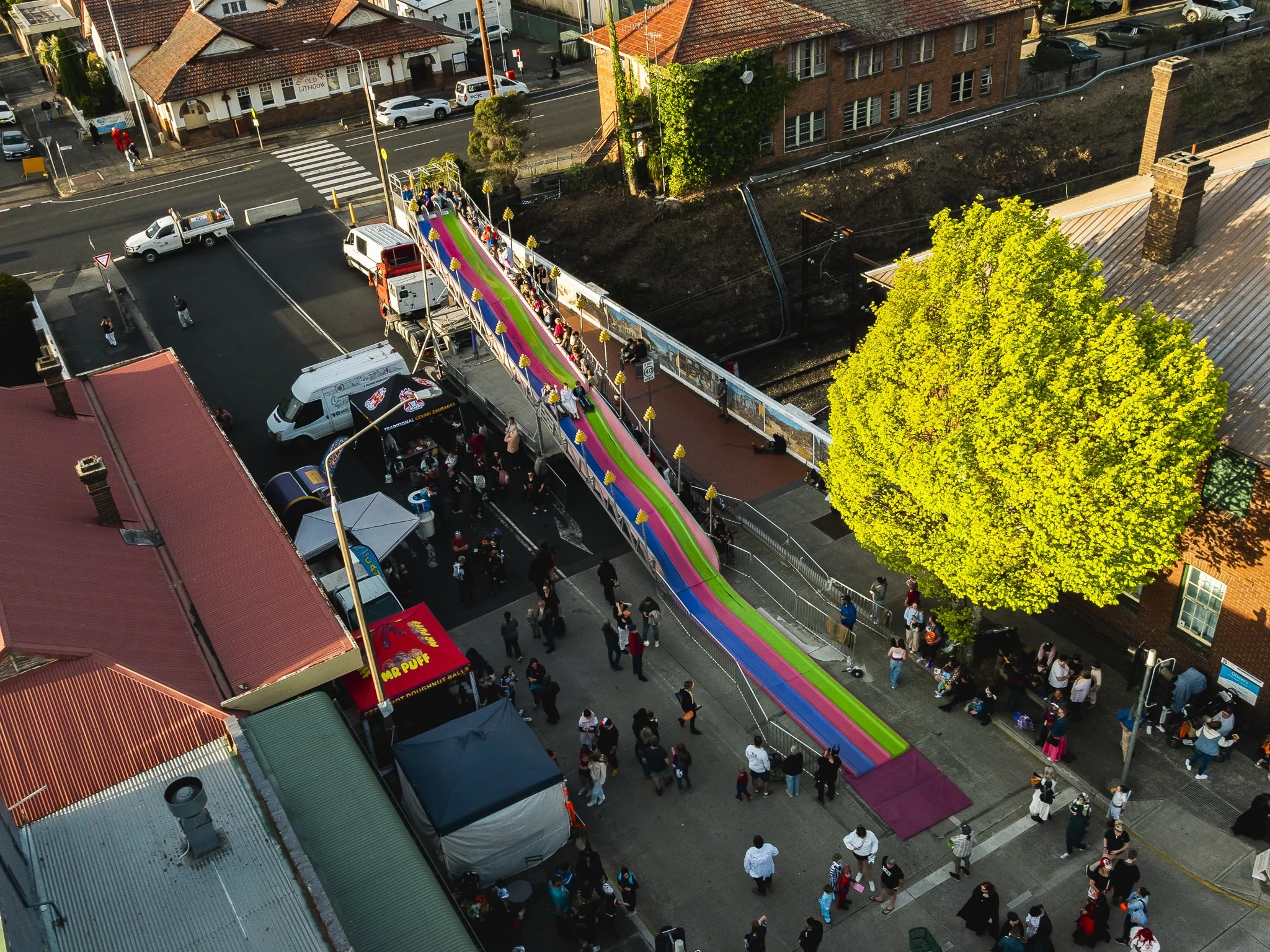 An aerial view of a colorful inflatable slide set up on a city sidewalk, with children and adults gathered around. There are tents and food trucks nearby, with a large green tree and brick buildings surrounding the area.