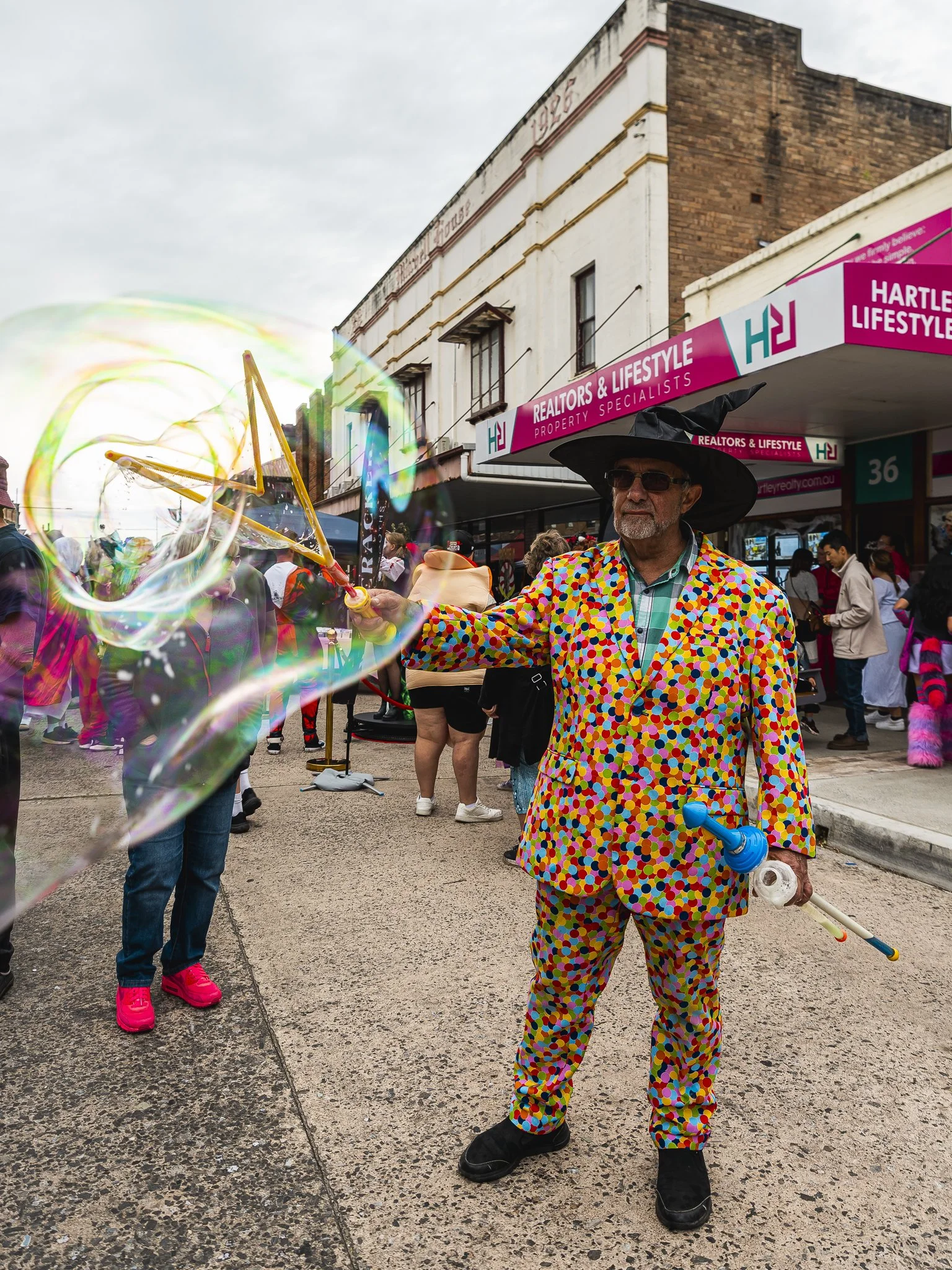 A man wearing a colorful polka dot suit and a large black hat is making large soap bubbles with a bubble wand in a street fair or festival. There are people around and storefronts in the background.