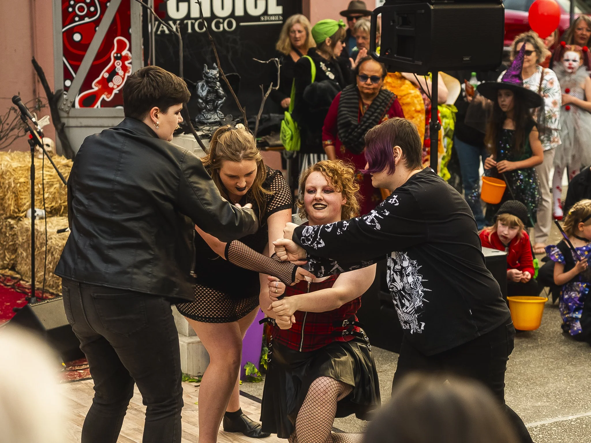 Three women in dark clothing appear to be engaged in a playful wrestling match while a crowd of children and adults watch in the background at an outdoor event or street festival.