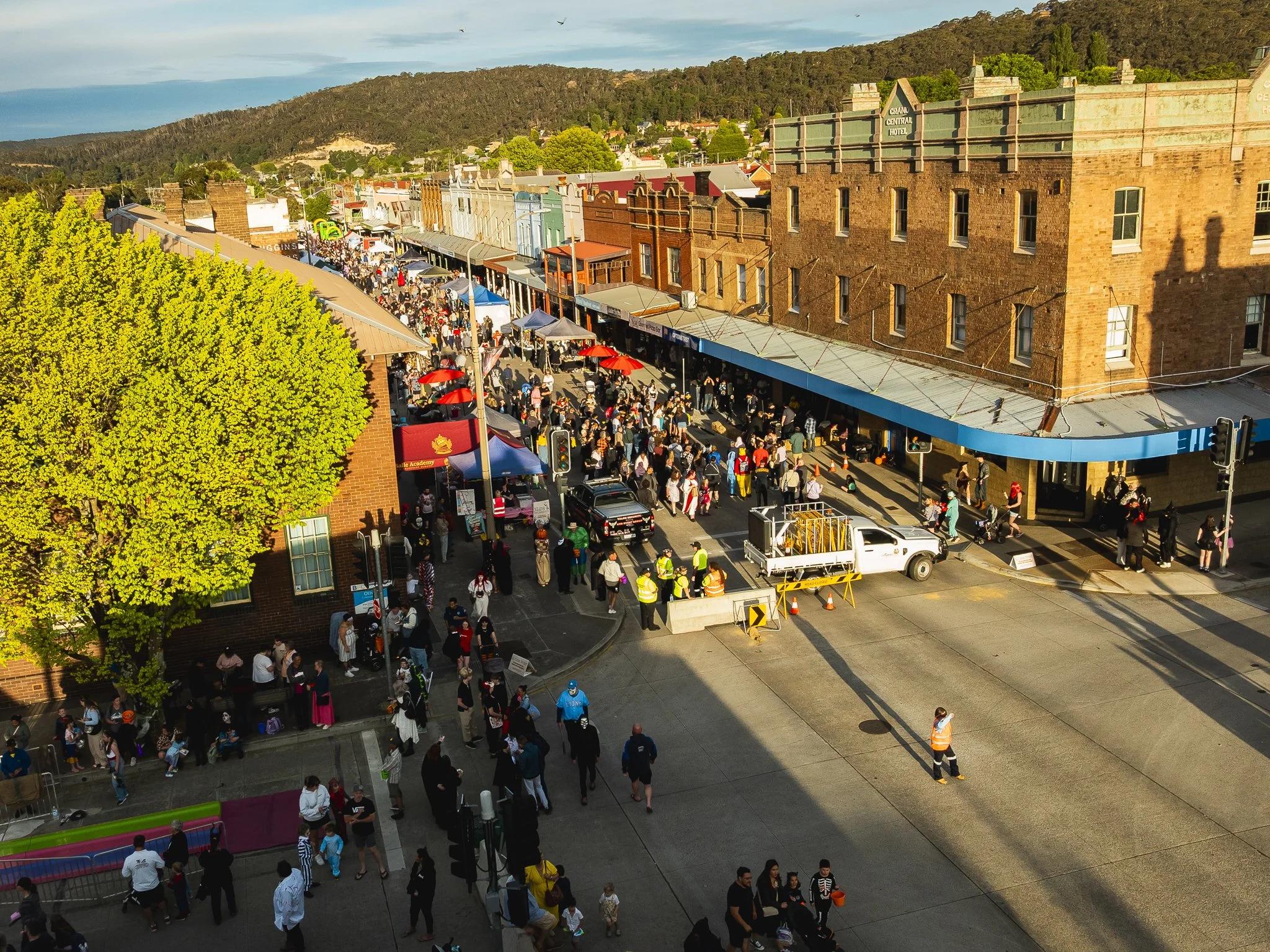 A busy downtown street with many pedestrians, vendors, and street performers during daytime. Crowded sidewalks, a large tree, and historic brick buildings with shops line the street. Some vehicles and construction workers are visible.