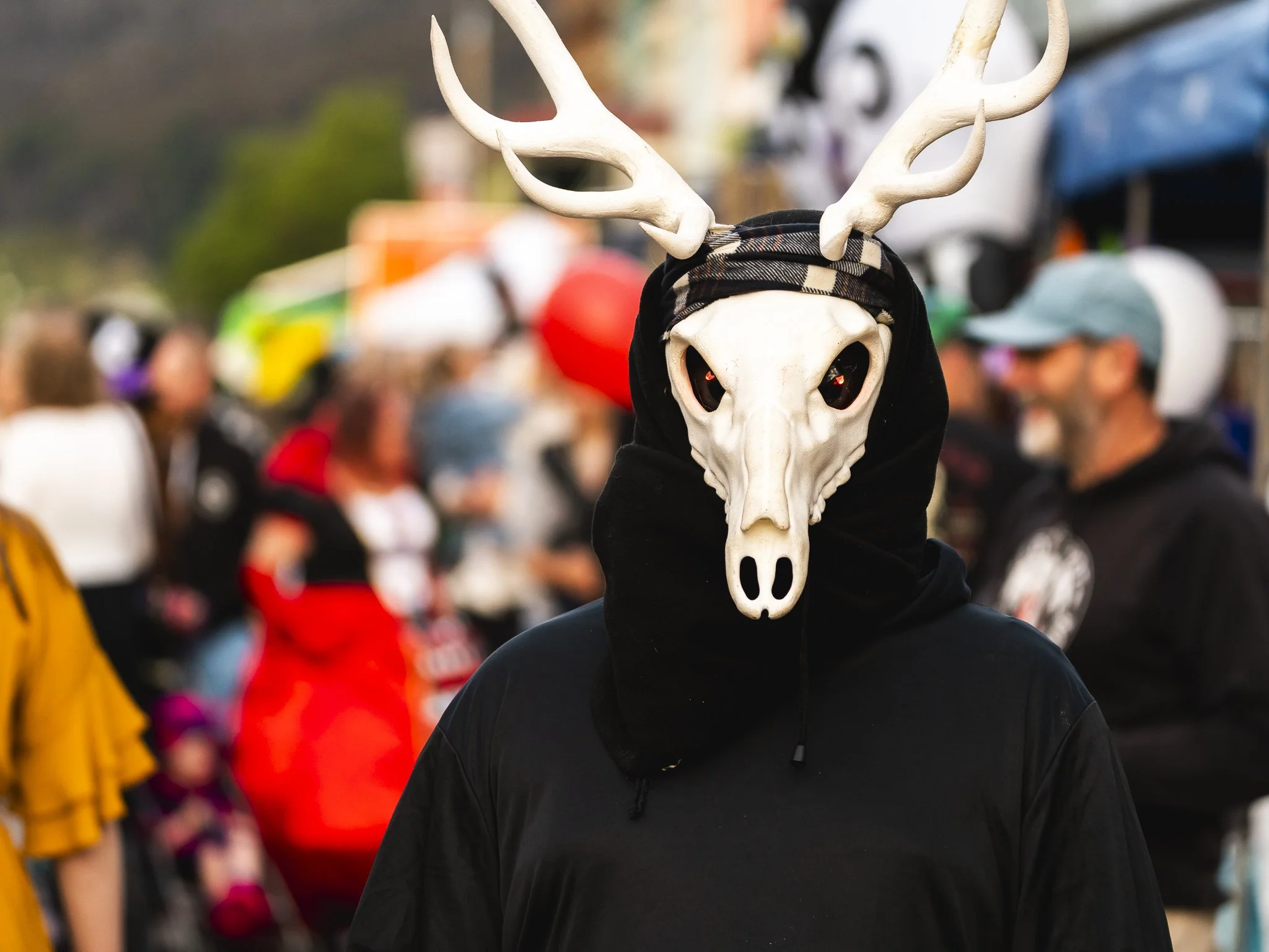 A person wearing a deer skull mask with antlers, black hoodie, and black jacket at an outdoor event with blurred crowd and tents in background.