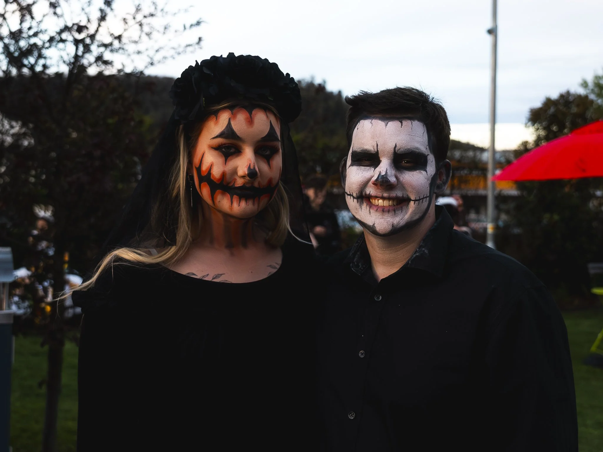 Two people dressed in Halloween costumes with skeleton and pumpkin face paint, standing outdoors during dusk.