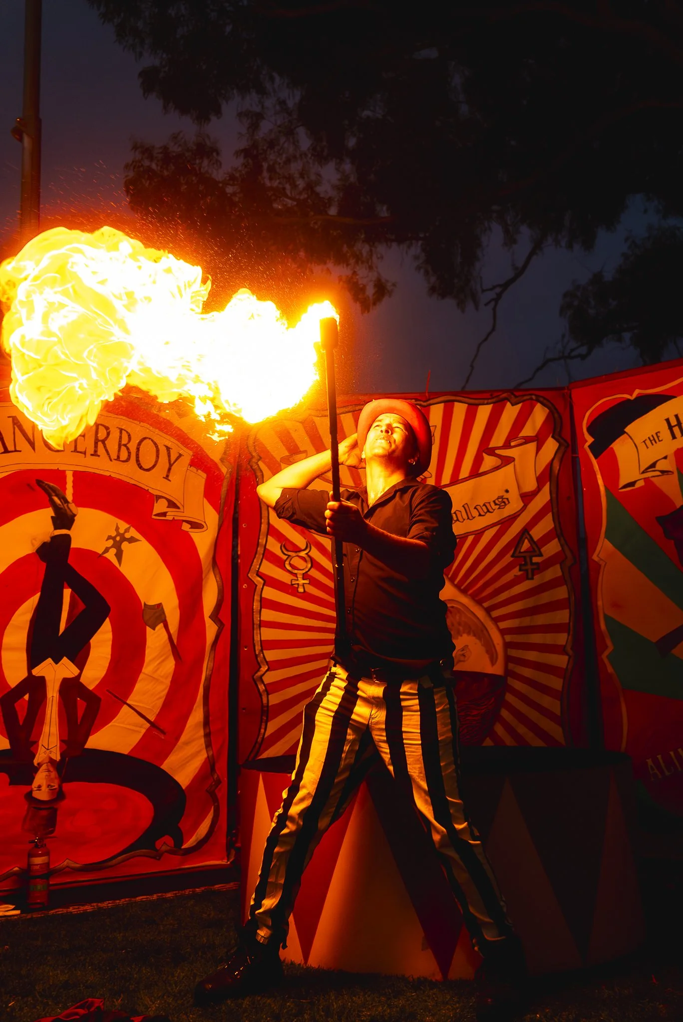 A performer in black and yellow striped pants, holding a torch and blowing fire during a circus act at night.