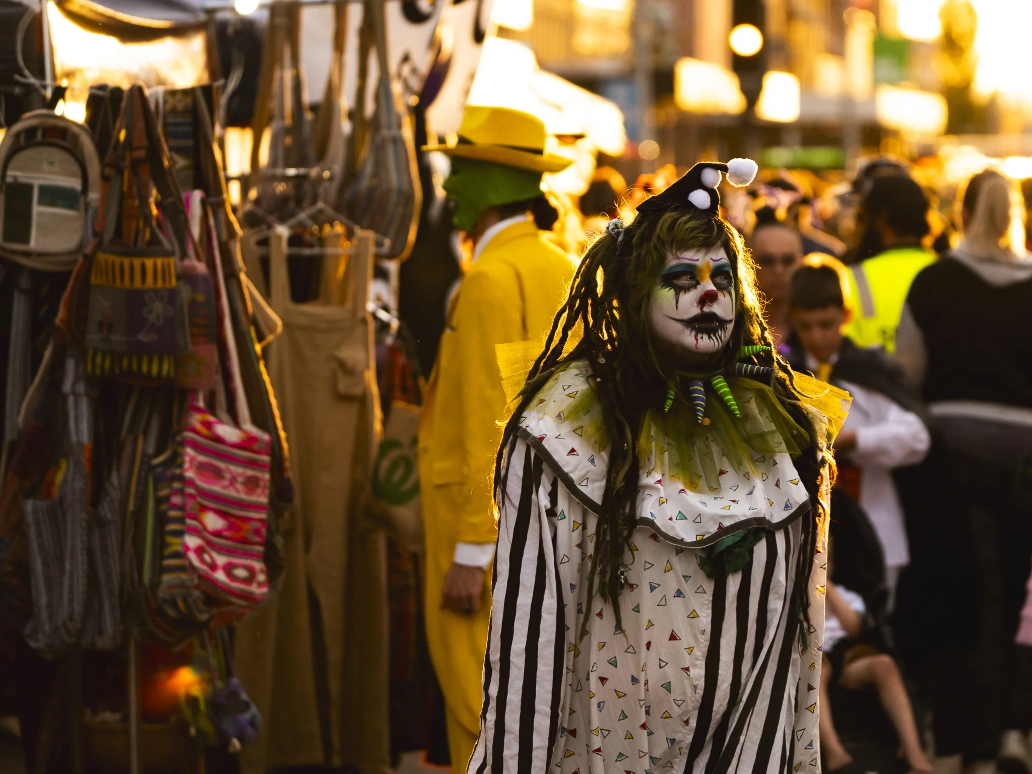 A person dressed as a clown with face paint and dreadlocks, wearing a striped costume and a collar, stands at a crowded outdoor market during sunset, with various people and vendor stalls in the background.