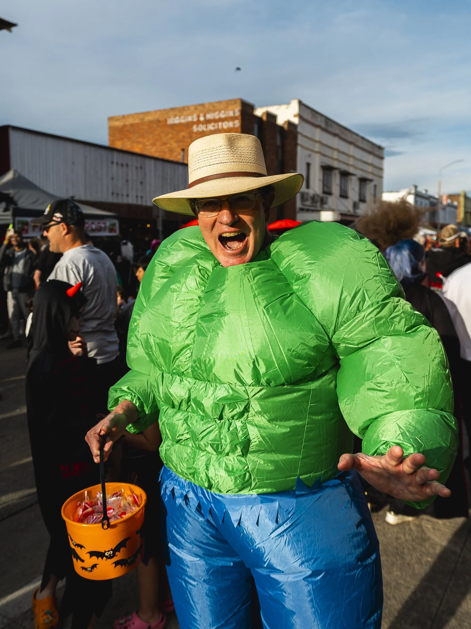 Person dressed in a green inflatable costume, wearing a straw hat and glasses, participating in a festive event outdoors.
