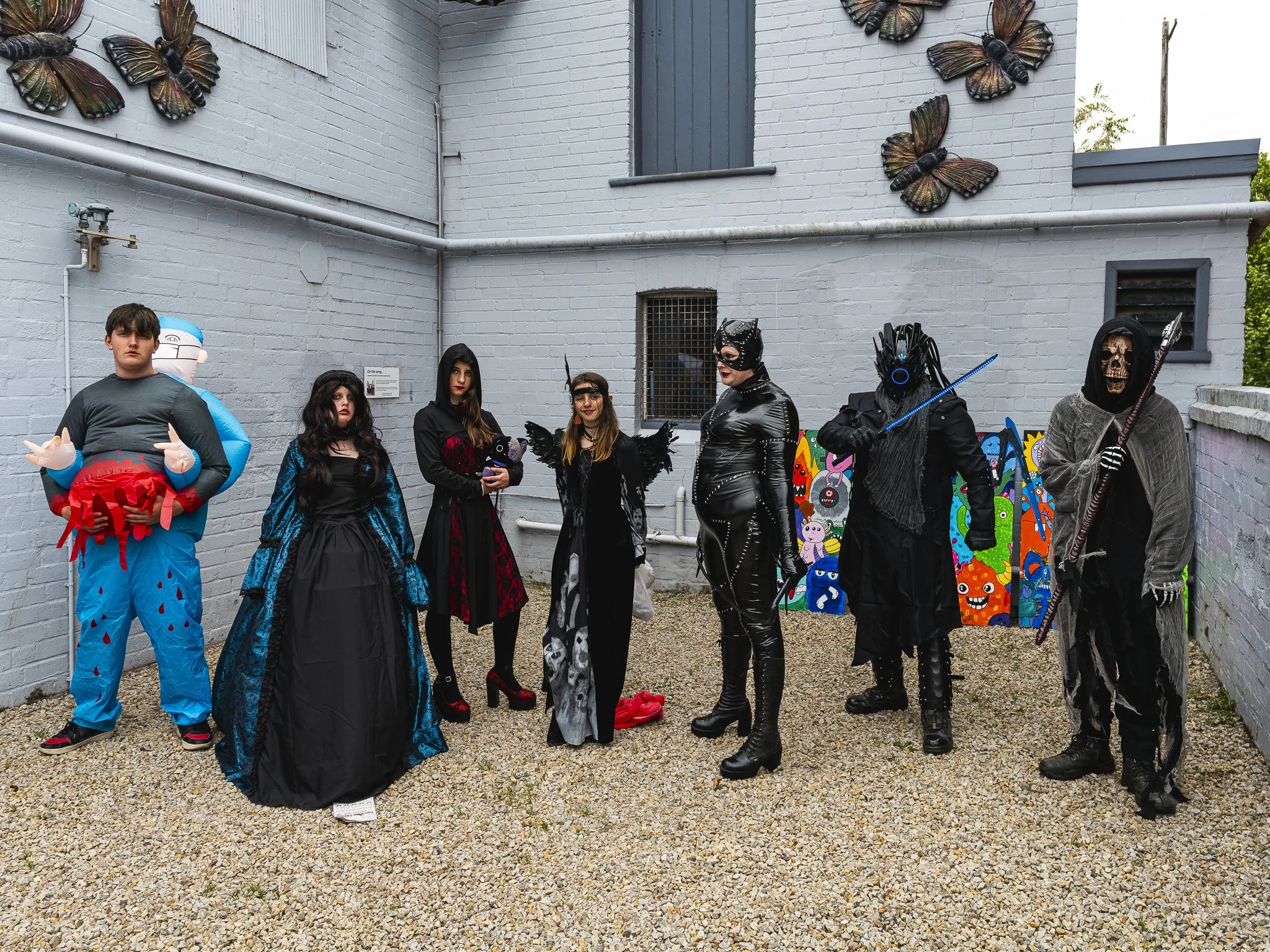 People dressed in Halloween costumes, including a person as a blue cartoon character with a crab, a woman in a blue and black dress, a woman in a red and black dress holding a small black object, a woman in skeleton print, a person in a shiny black o