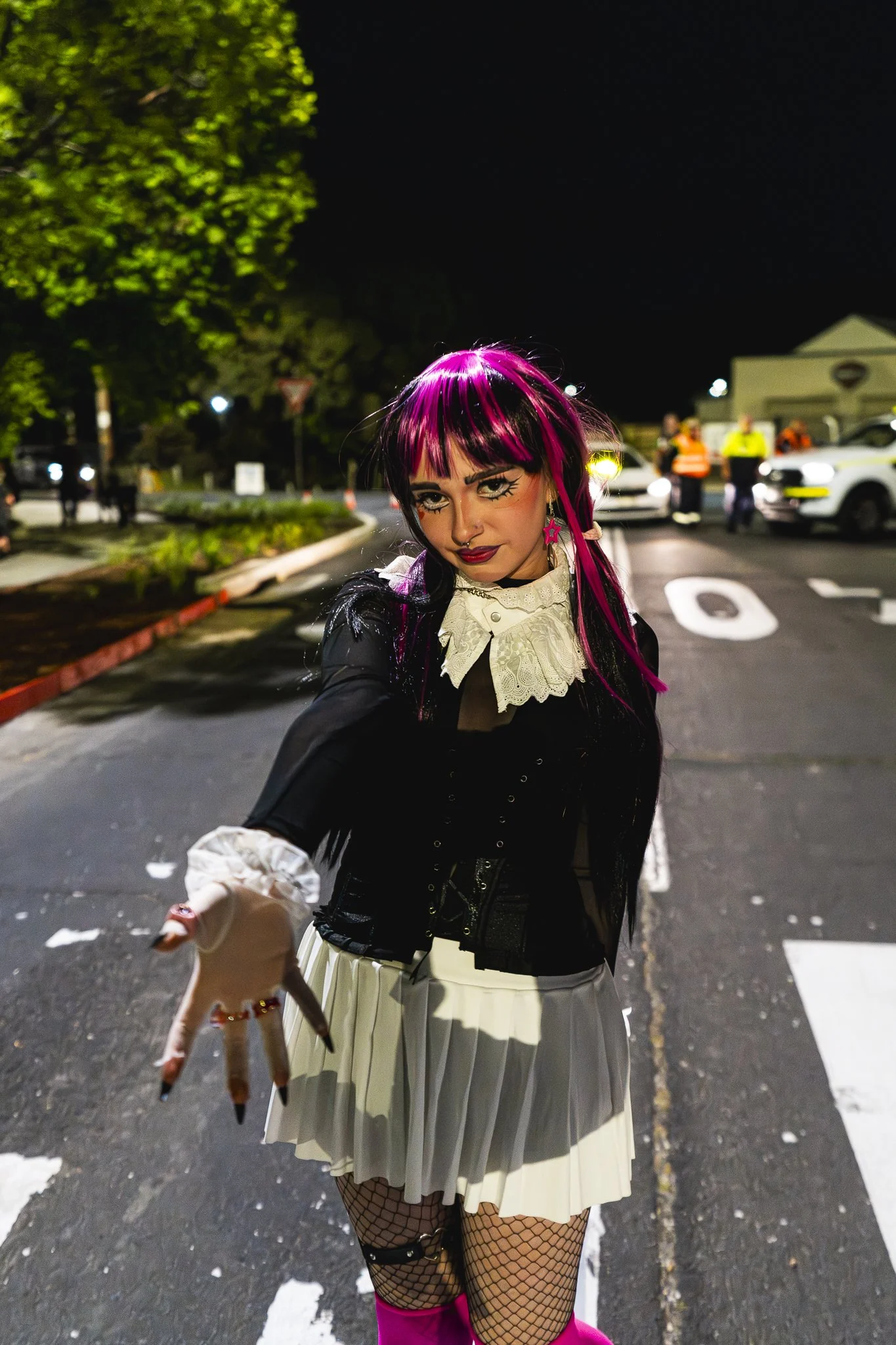 A woman with pink and black hair reaching out towards the camera at night on a city street, with police officers and cars in the background.