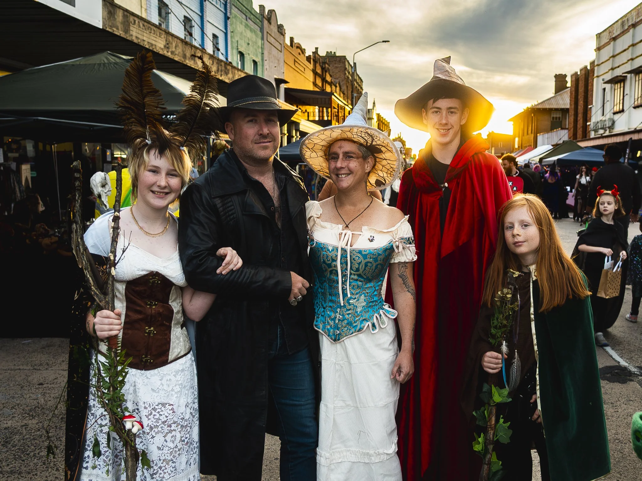 A group of five people dressed in fantasy and witch costumes standing together at an outdoor event during sunset, with a lively street market in the background.