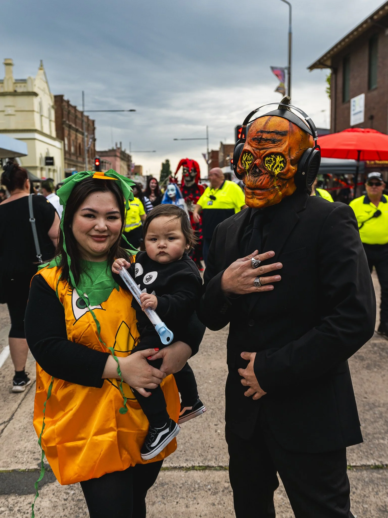 A woman holding a young child at a street event, with a person in a skull mask wearing headphones to her right, and other costumed people and police officers in the background.