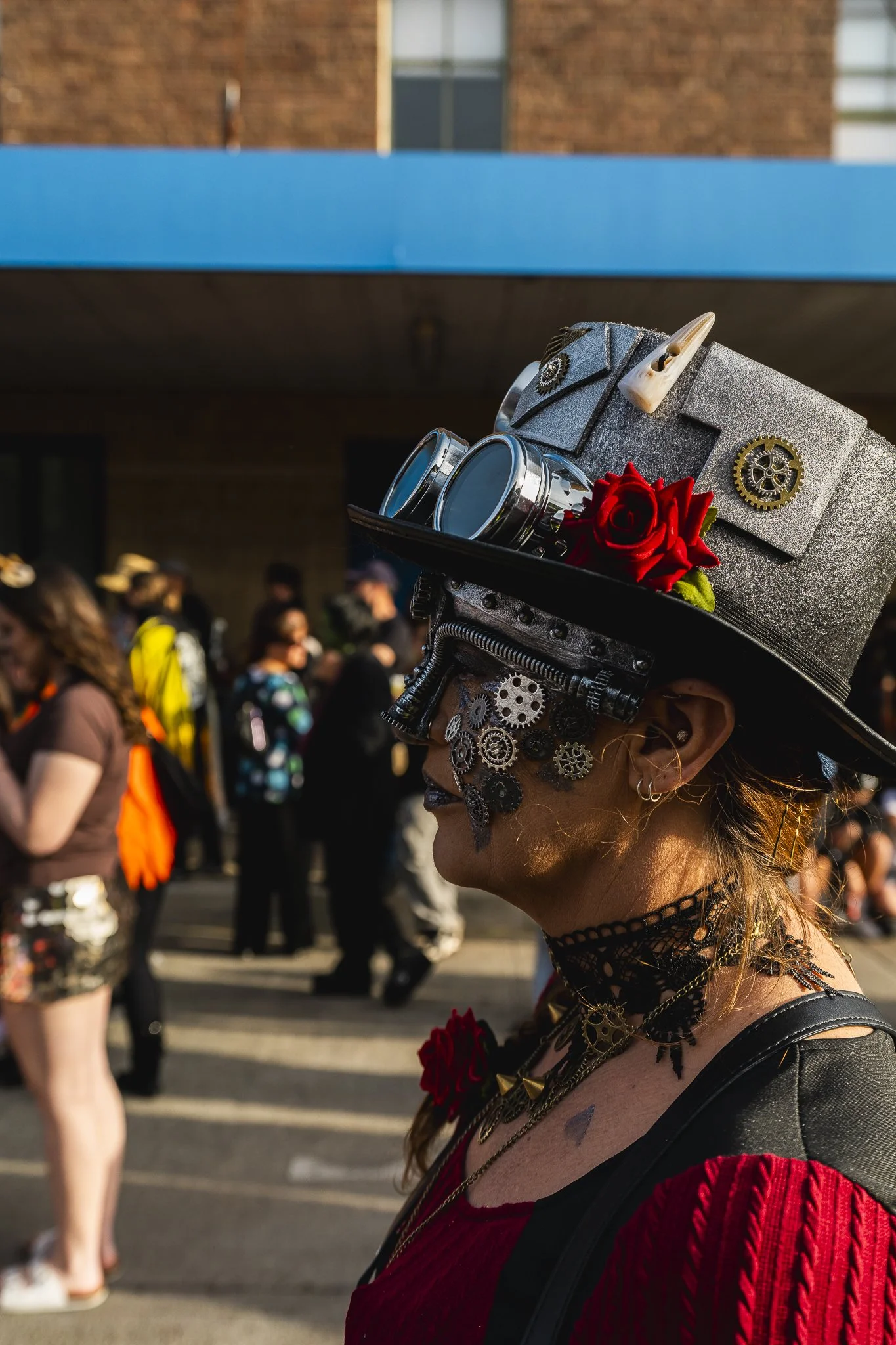A woman wearing a steampunk-inspired outfit, including a large gray top hat decorated with gears, a red rose, and goggles, is part of a crowd in an outdoor urban setting.