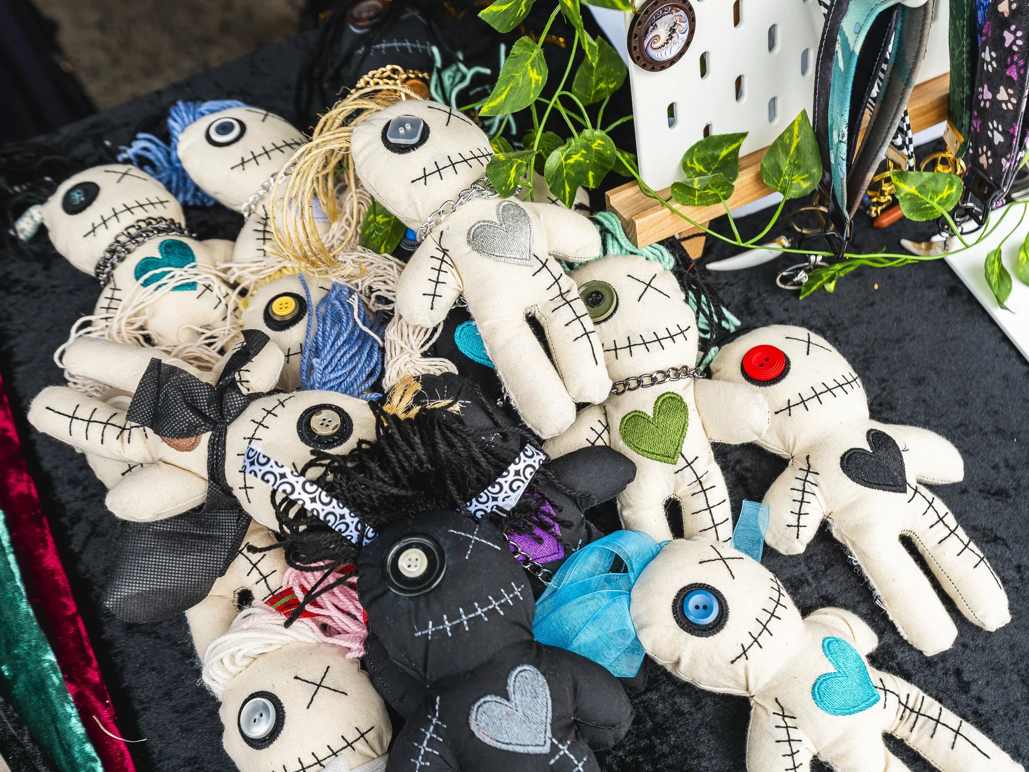 A collection of handmade fabric dolls resembling skeletons with stitched eyes and mouths, featuring colorful hearts, buttons, and yarn details, displayed on a dark surface with some plant leaves and accessories nearby.