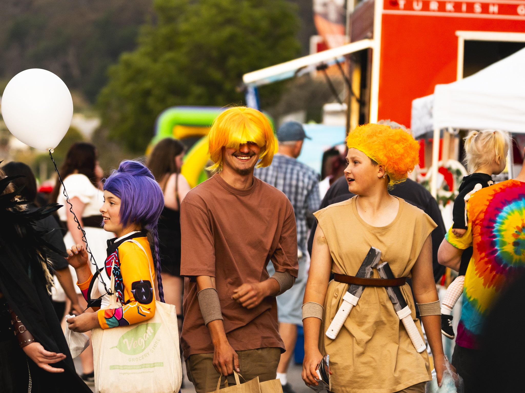 People in colorful costumes and wigs at an outdoor festival, with tents and booths in the background.