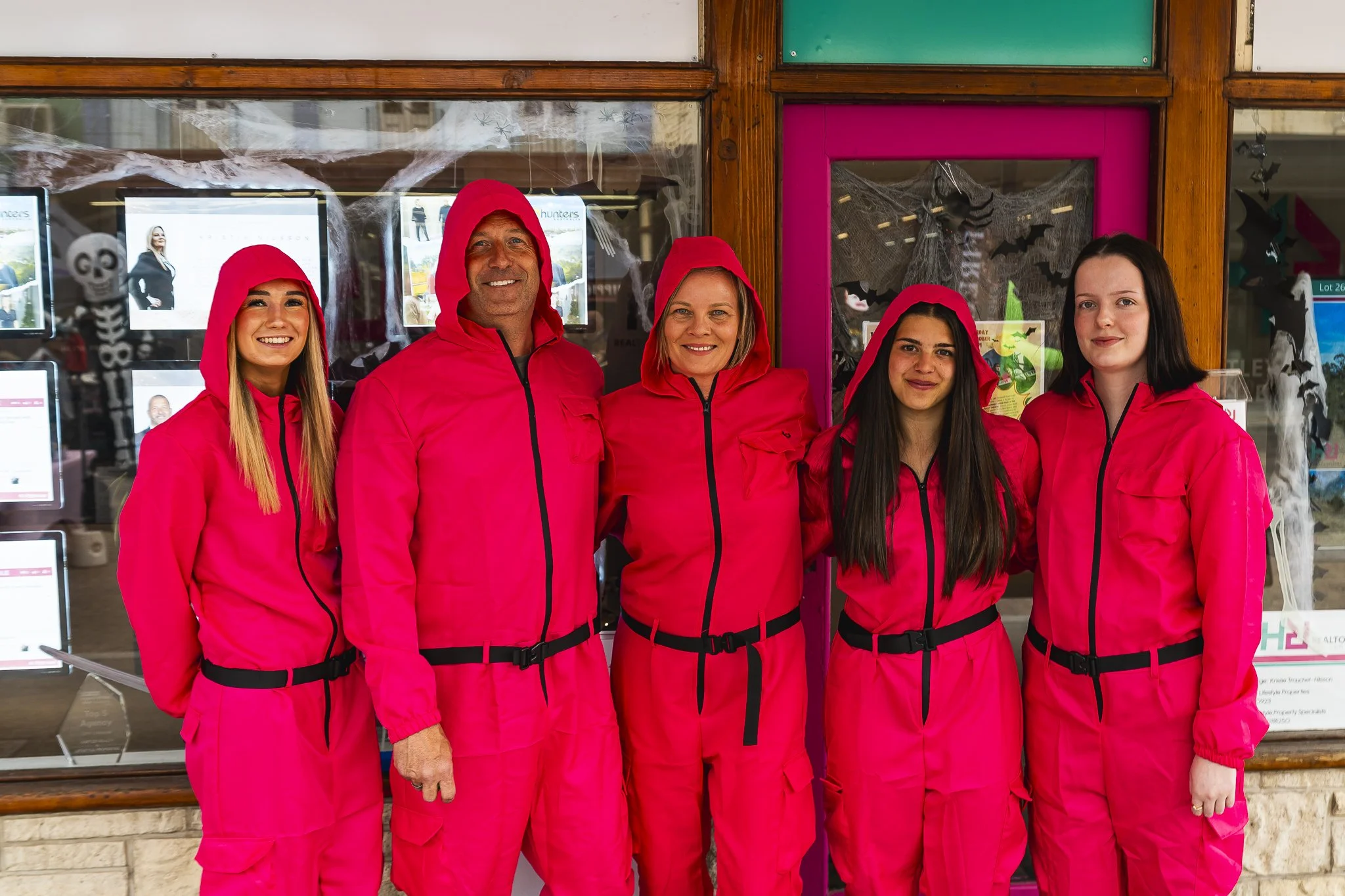Group of five people dressed in bright pink hooded jumpsuits, standing outside a building with Halloween decorations.