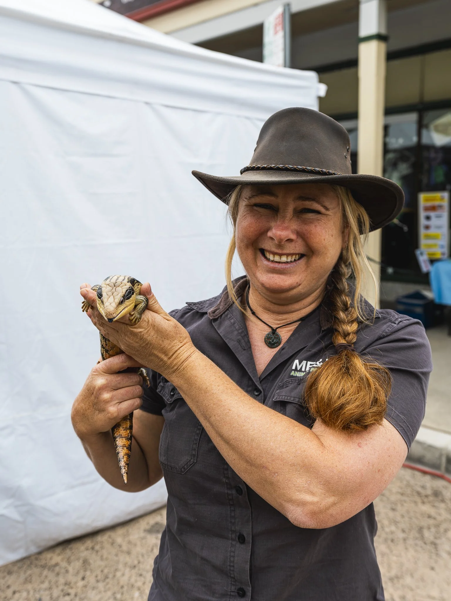 A woman wearing a cowboy hat, smiling, holds a large reptile, possibly a lizard, with a patterned skin. She is outdoors near a white tent or canopy.