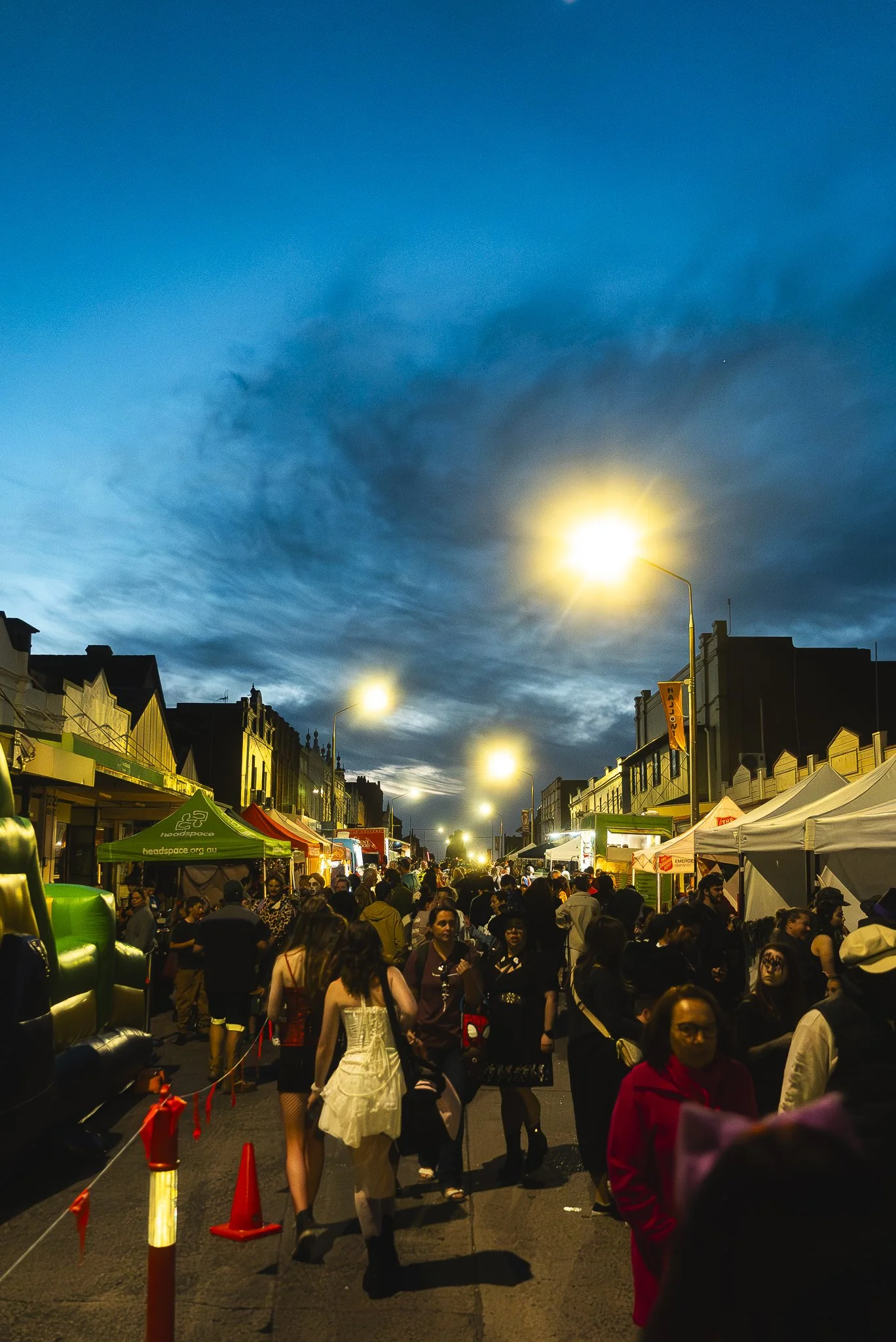 Crowd of people walking through a street market at dusk, with food stalls and tents on both sides and streetlights illuminating the scene.