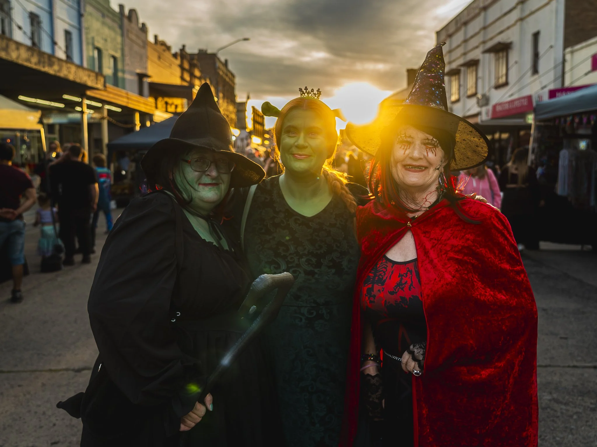 Three people dressed as witches and a witch in costume at a Halloween event during sunset on a busy street.