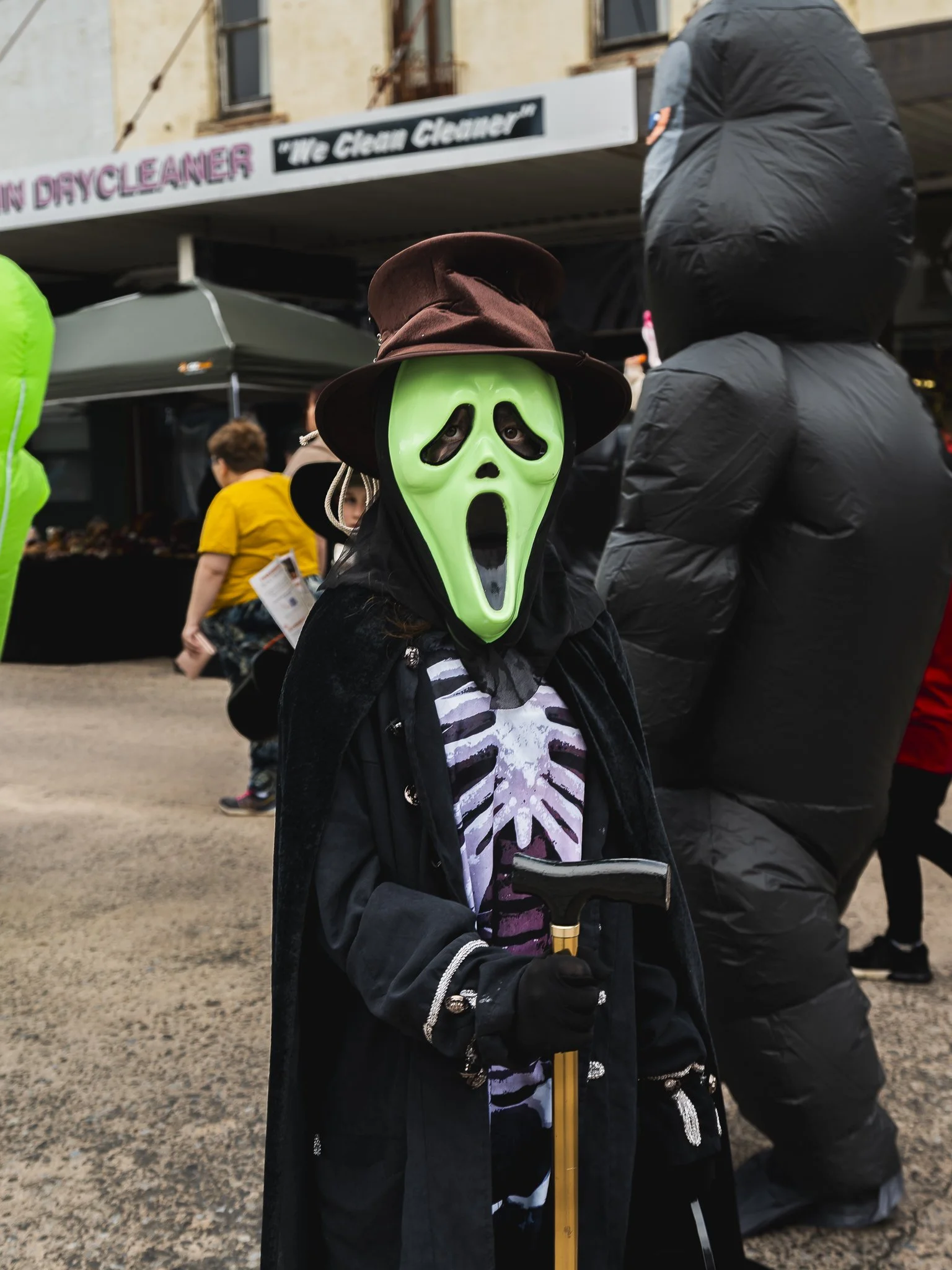 Person dressed in a skeleton costume with a green ghost face mask, wearing a brown hat, black cape, and gloves, holding a walking cane at a street event.
