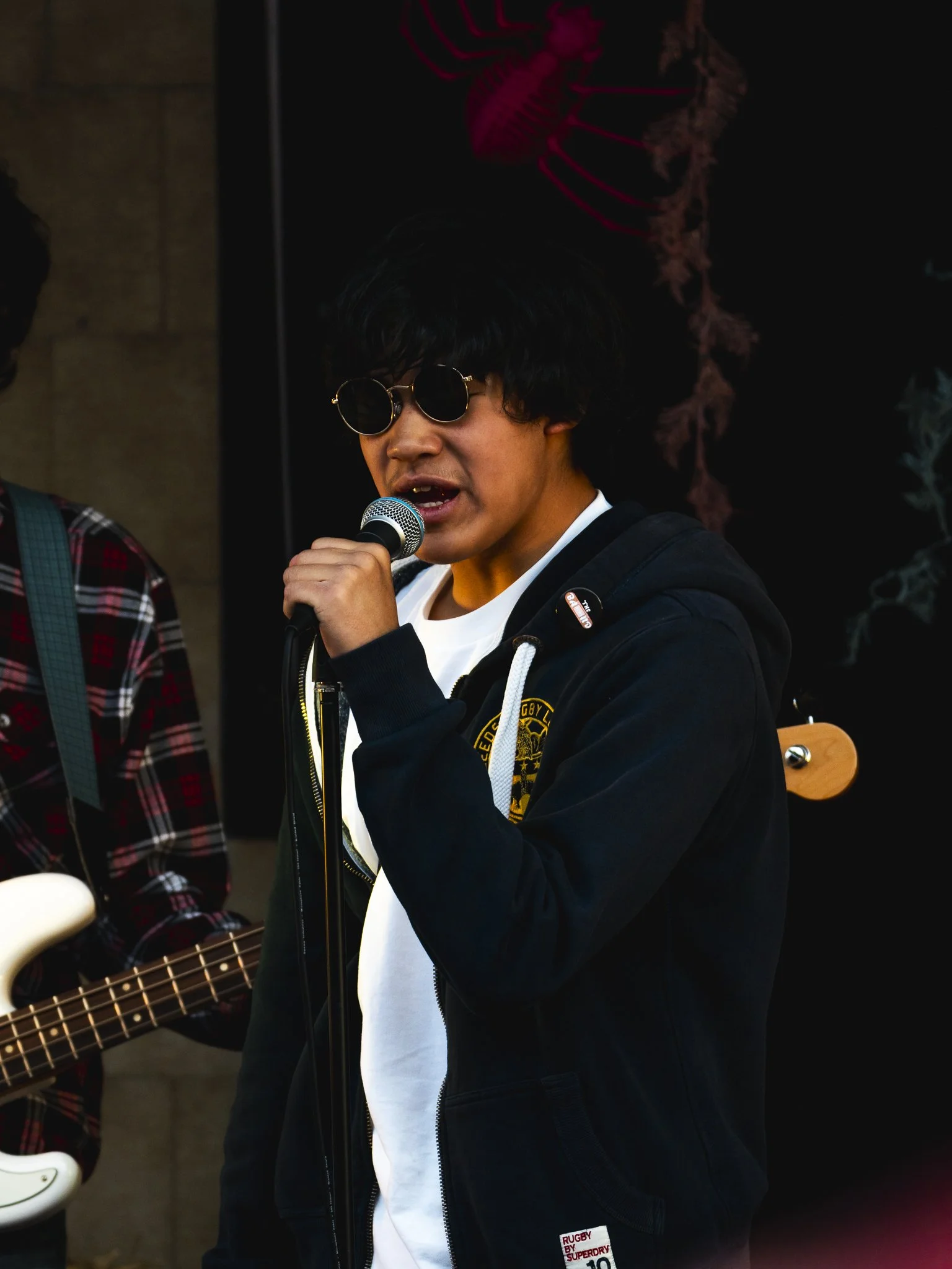 A young man with black curly hair and round dark sunglasses singing into a microphone, wearing a white shirt and a black hoodie, with a musician playing a bass guitar in the background.