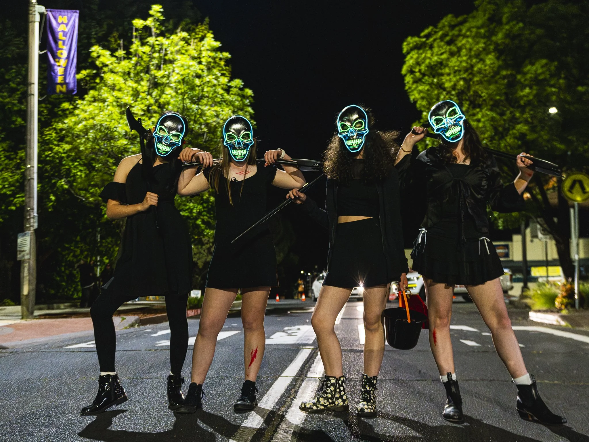 Four women in dark clothing, wearing glowing skull masks, carrying weapons and a bucket, standing in the middle of a street at night, with Halloween decorations and street signs in the background.