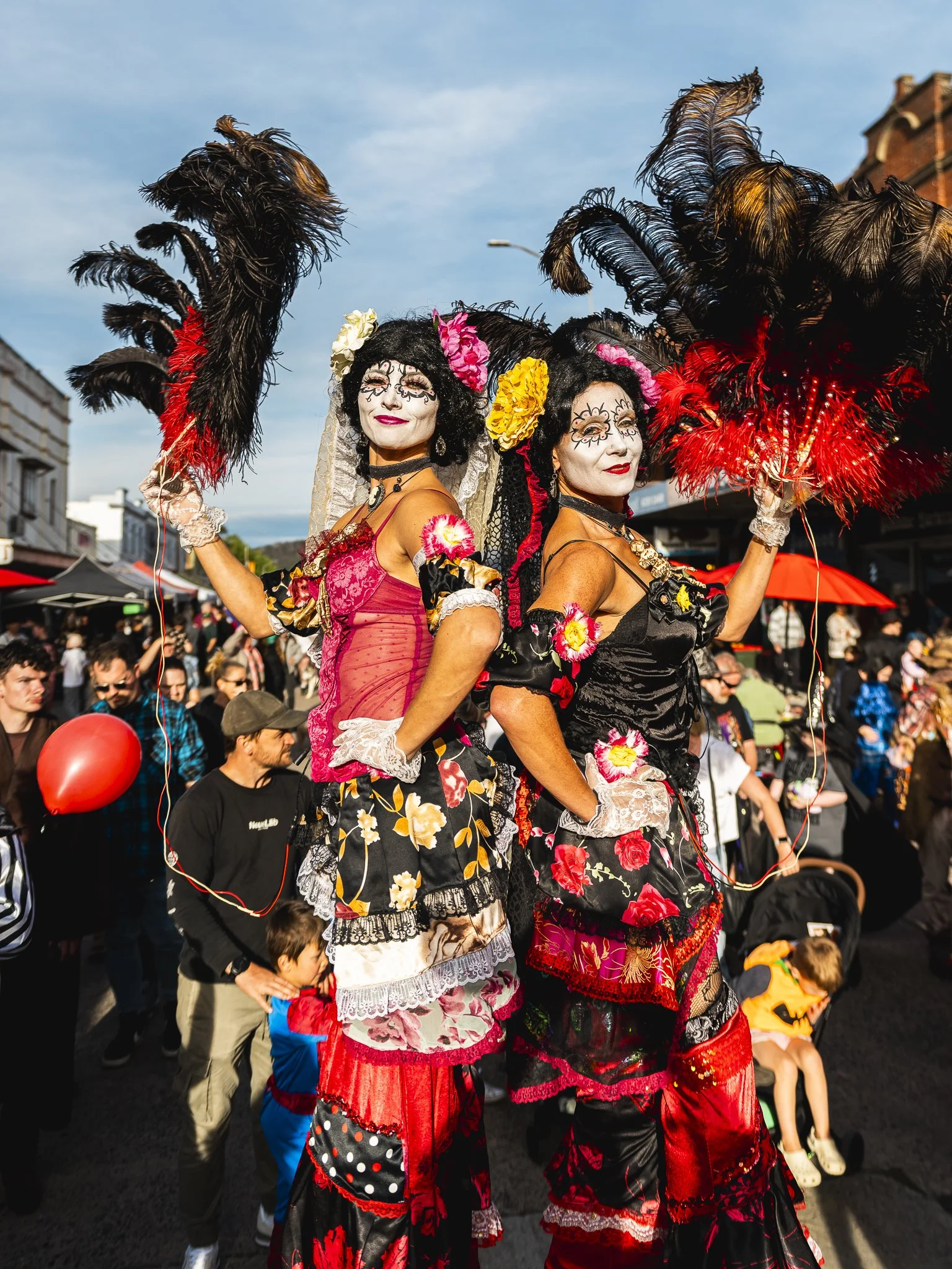 Two performers dressed as Mariachi musicians in colorful, elaborate costumes with large feathered headdresses, standing on stilts during a street festival with crowd in the background.